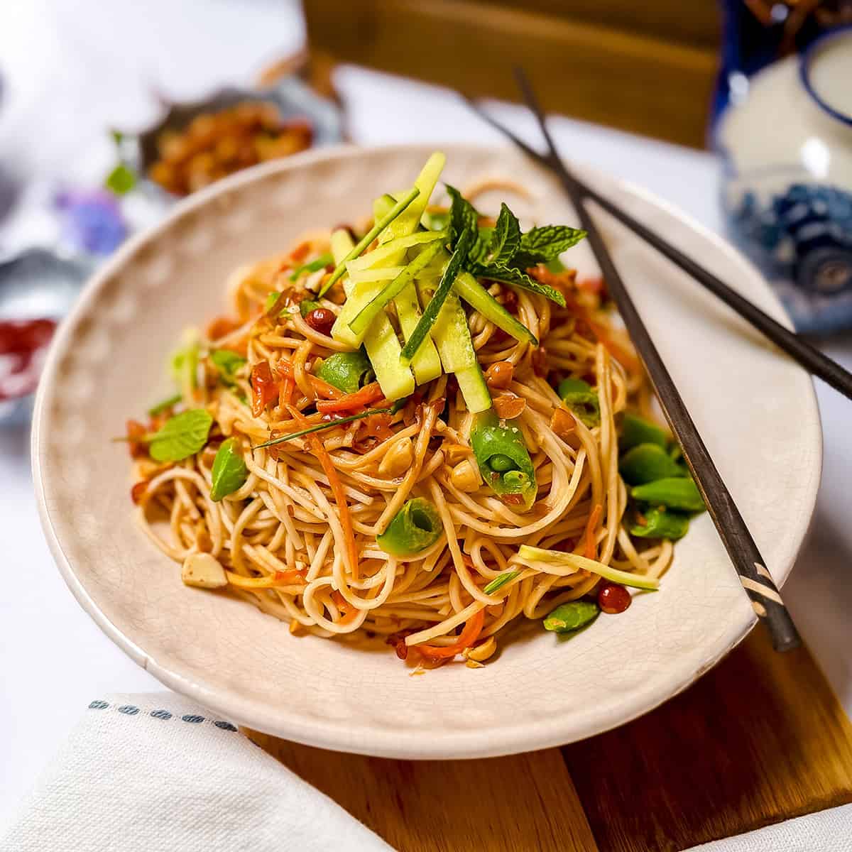 A plate of Cold Peanut Sesame Noodles with sliced vegetables and herbs, garnished with cucumber and mint, with chopsticks resting on the plate.