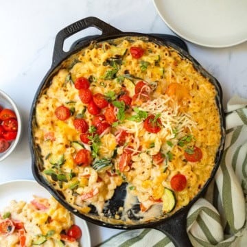 A cast iron skillet filled with baked Skillet Mac and Cheese, topped with cherry tomatoes, fresh herbs, and shredded cheese, sits on a marble surface next to plates and a striped napkin.
