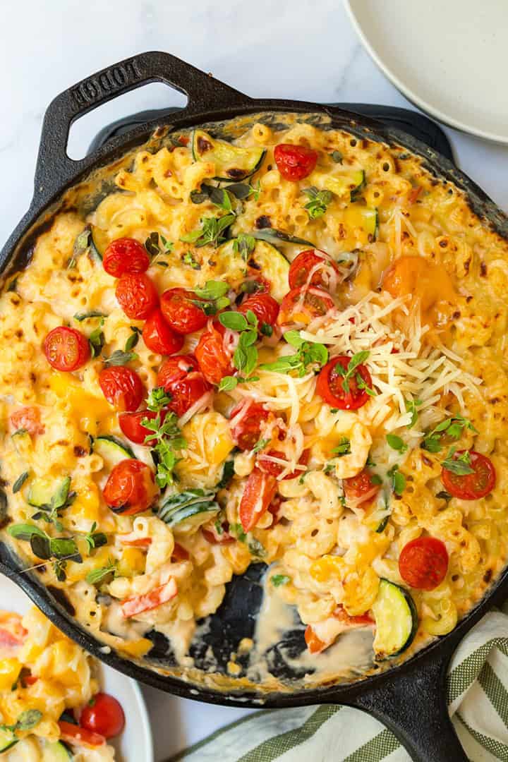 A cast iron skillet filled with baked Skillet Mac and Cheese, topped with cherry tomatoes, fresh herbs, and shredded cheese, sits on a marble surface next to plates and a striped napkin.