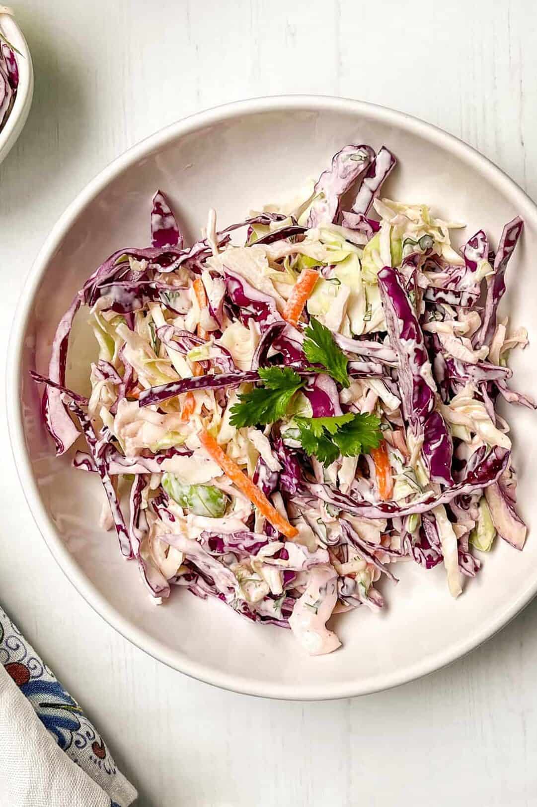 A bowl of coleslaw with shredded red and green cabbage, carrots, and creamy dressing, garnished with parsley, on a light surface-perfect as a refreshing side for salmon tacos.