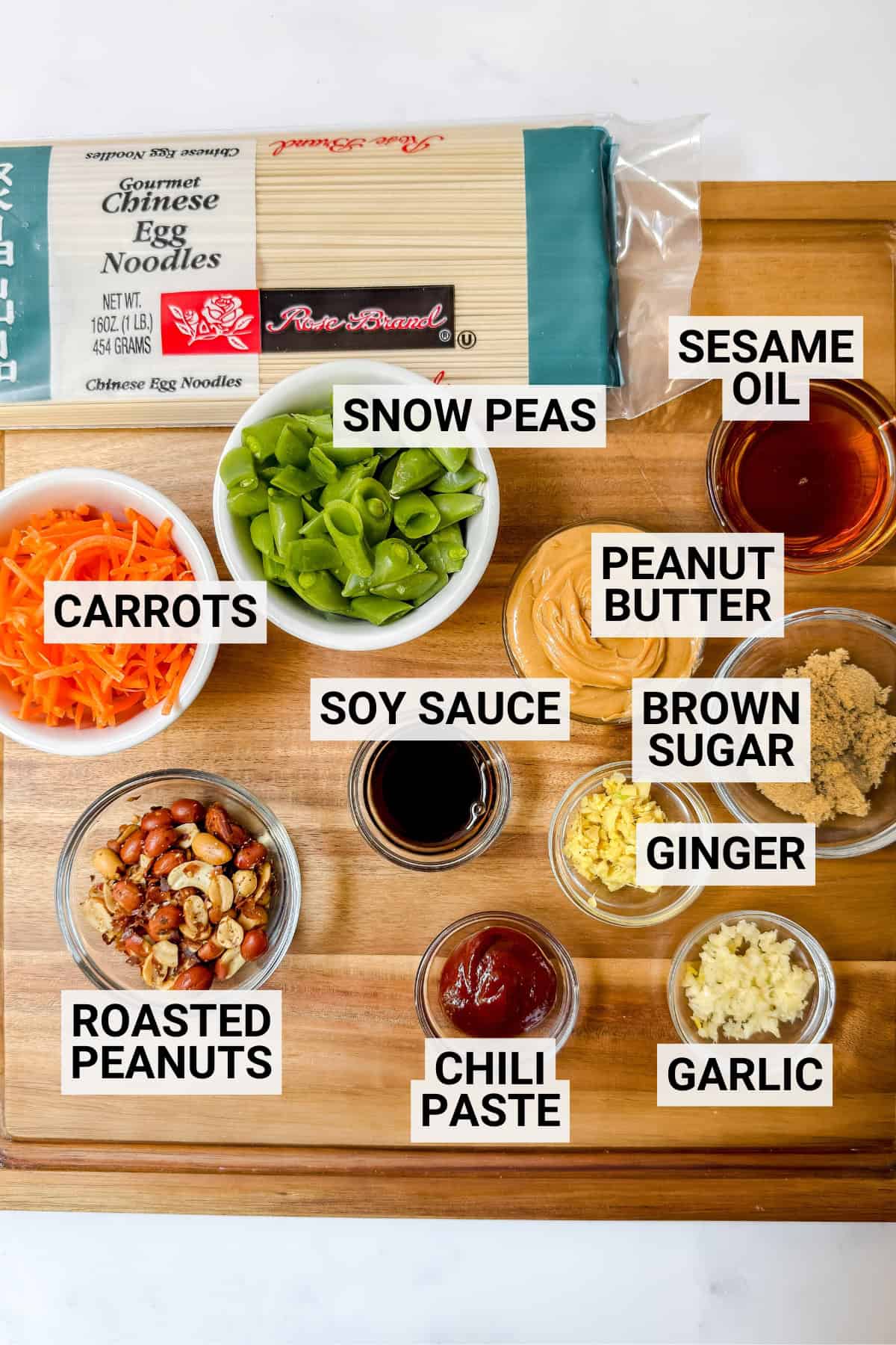A wooden board with labeled ingredients for Cold Peanut Sesame Noodles: Chinese egg noodles, snow peas, carrots, soy sauce, roasted peanuts, chili paste, ginger, garlic, brown sugar, peanut butter, and sesame oil.