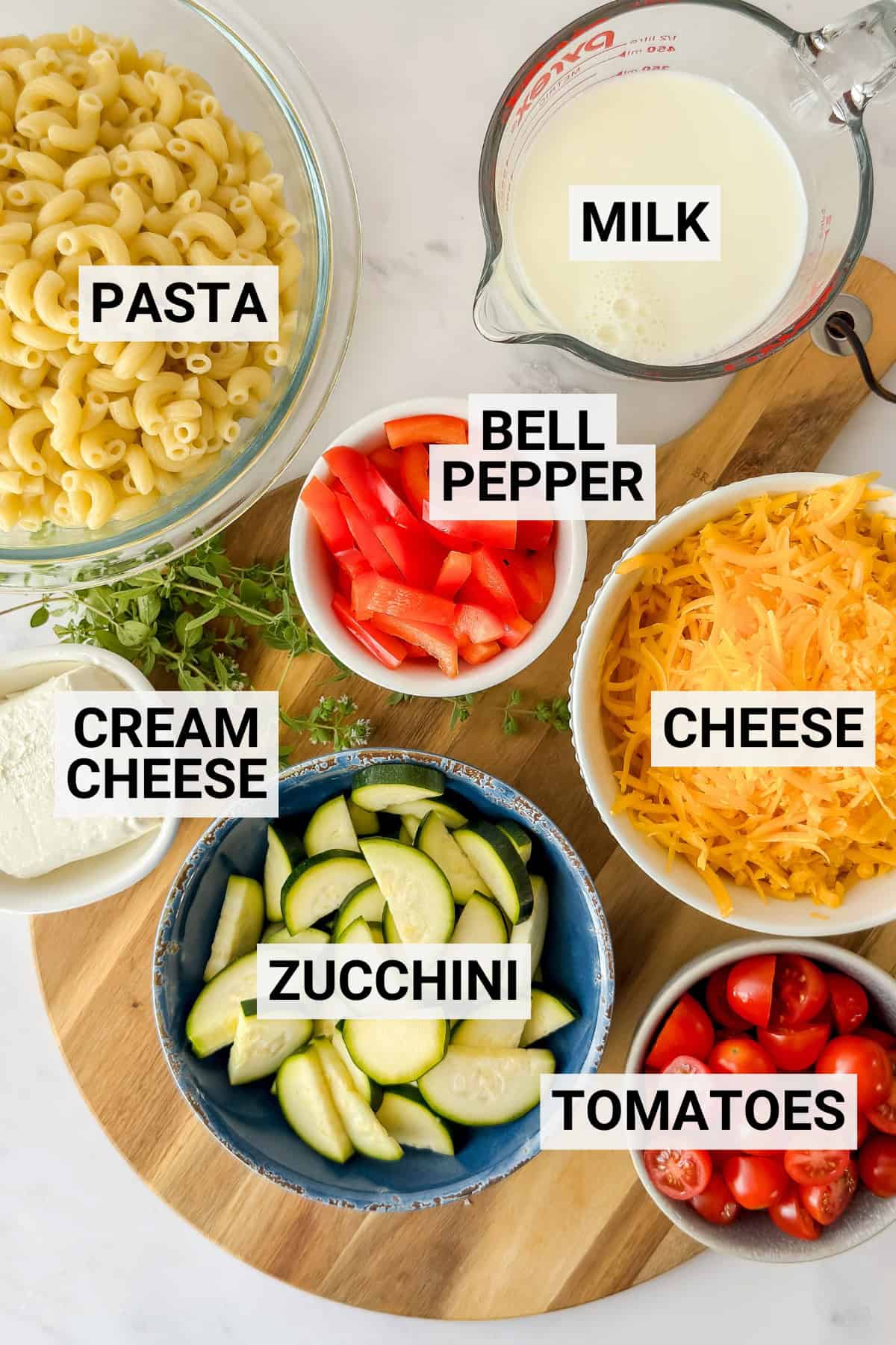 Bowls of uncooked macaroni, sliced zucchini, chopped bell pepper, cherry tomatoes, shredded cheese, cream cheese, and a measuring cup of milk for making skillet macaroni and cheese.