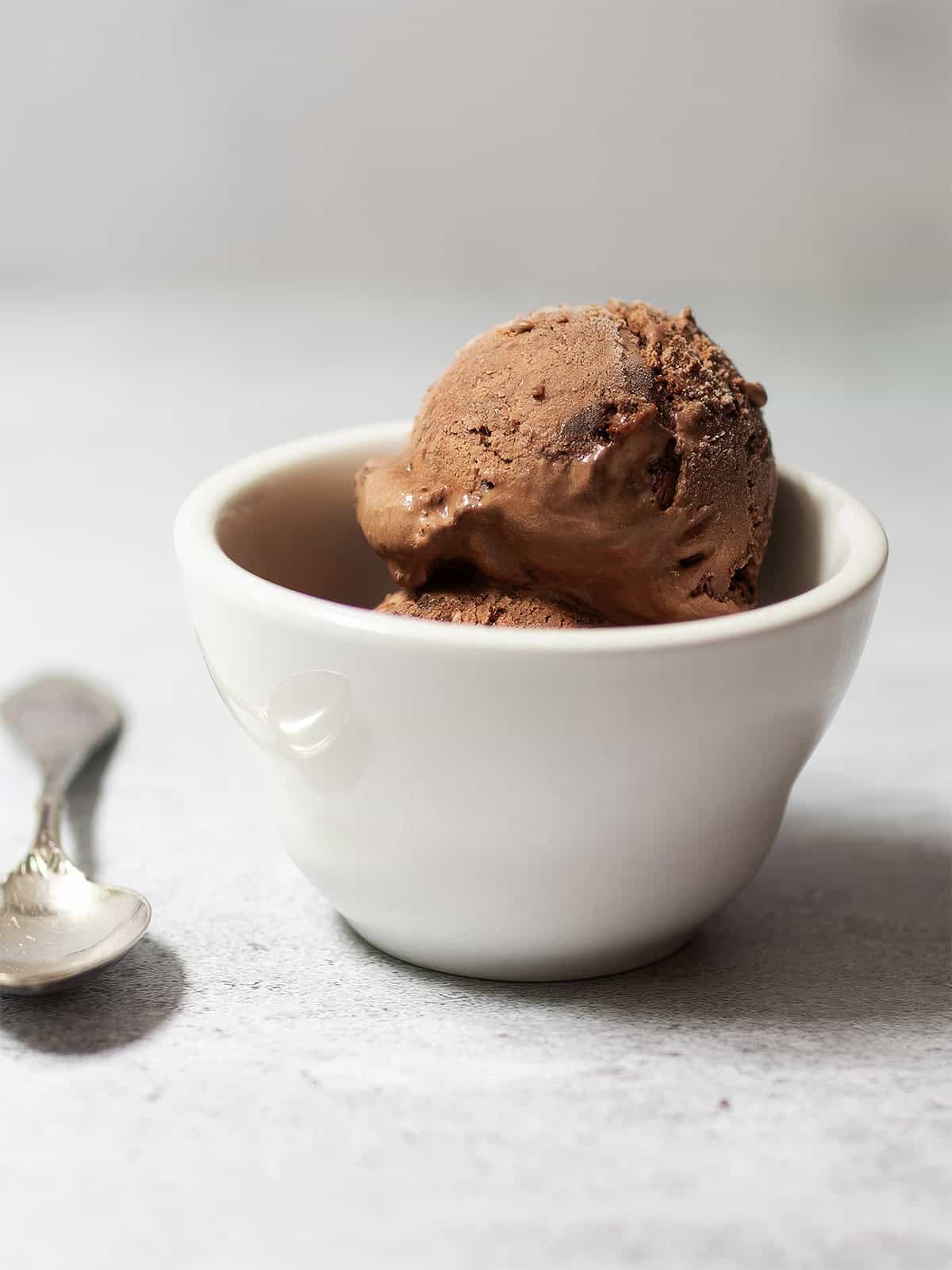 Two scoops of homemade no-churn chocolate ice cream in a white ceramic bowl beside a silver spoon.