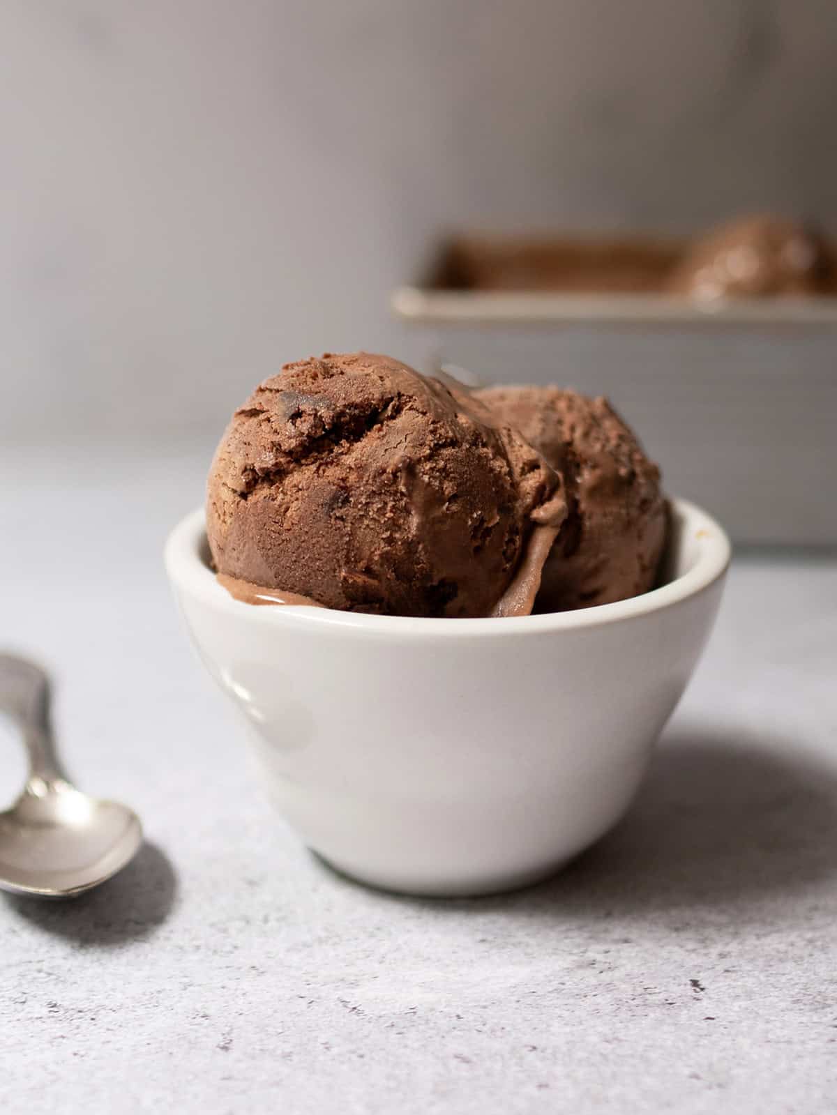 Two scoops of homemade no-churn chocolate ice cream in a white bowl, with a spoon and loaf pan in the background.