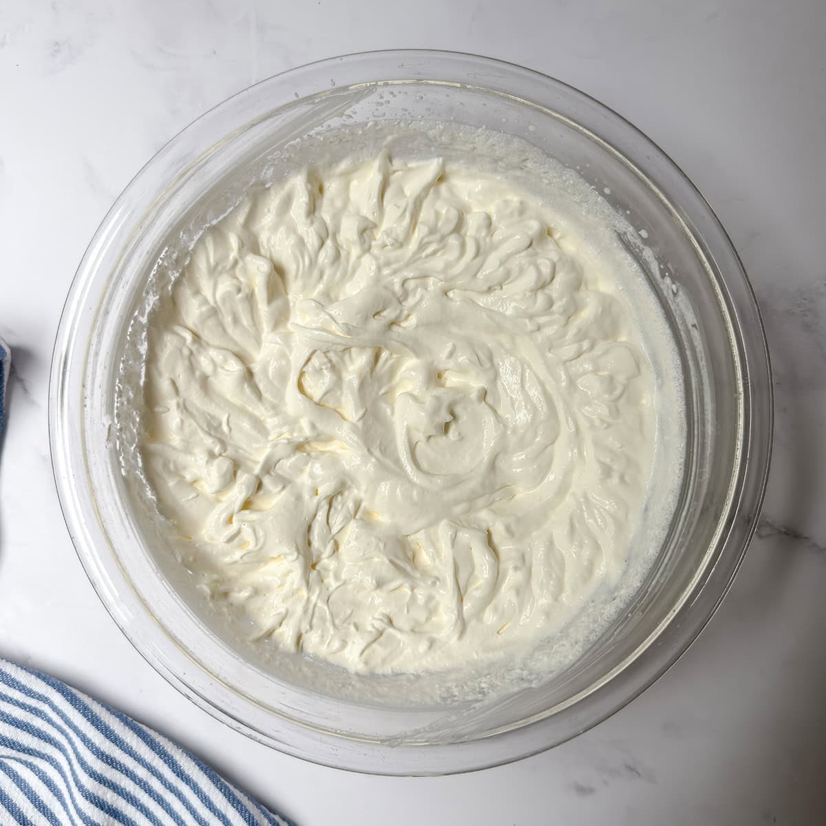 Heavy cream whipped to firm peaks in a glass bowl beside a blue-and-white striped cloth.