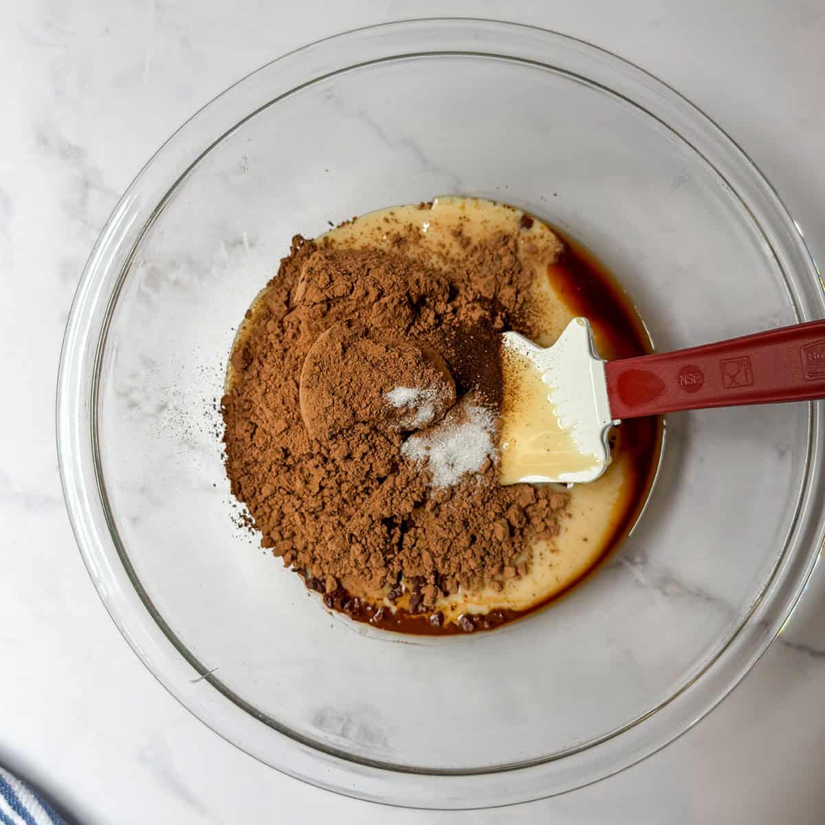 Sweetened condensed milk, cocoa powder, vanilla, espresso powder, and salt in a glass bowl with a red spatula.
