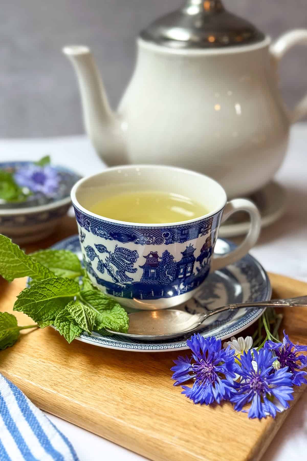 A cup of Liberty Tea in a blue and white patterned cup sits on a saucer with fresh herbs and blue flowers, on a wooden tray next to a teapot.