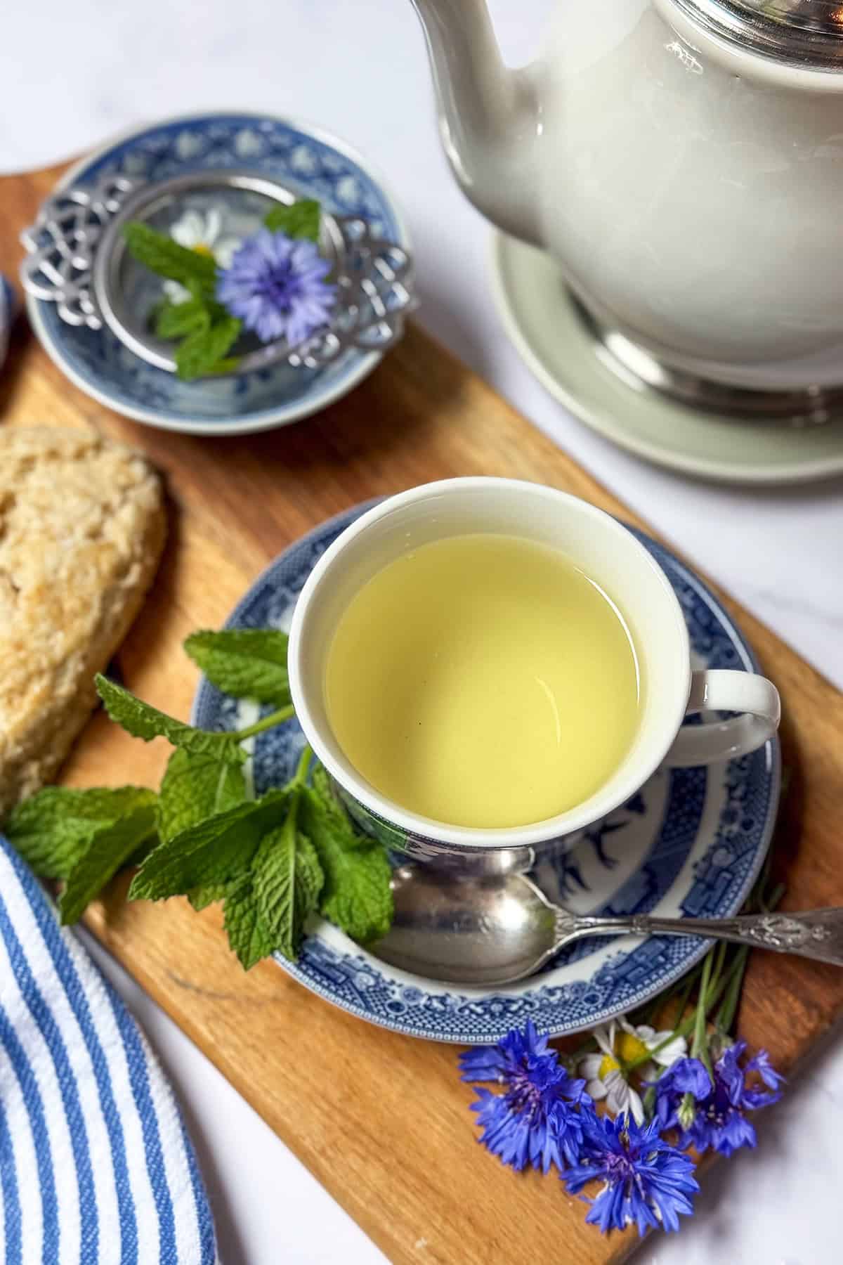 A cup of Liberty Tea on a blue and white saucer with fresh mint, a teaspoon, blue flowers, fresh herbs, and a biscuit on a wooden board next to a white teapot.