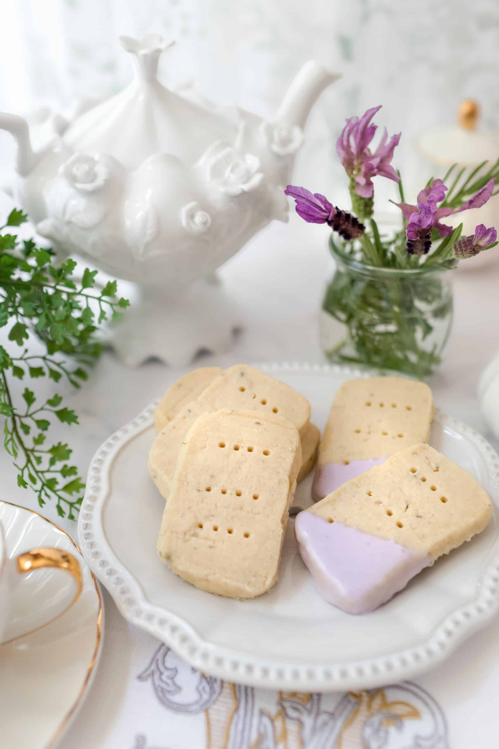 A plate of shortbread cookies, some dipped in peach and lavender icing, sits on a table with a teapot, a teacup, a small vase of purple flowers, and a green plant-perfect for an August Afternoon Tea in the garden.