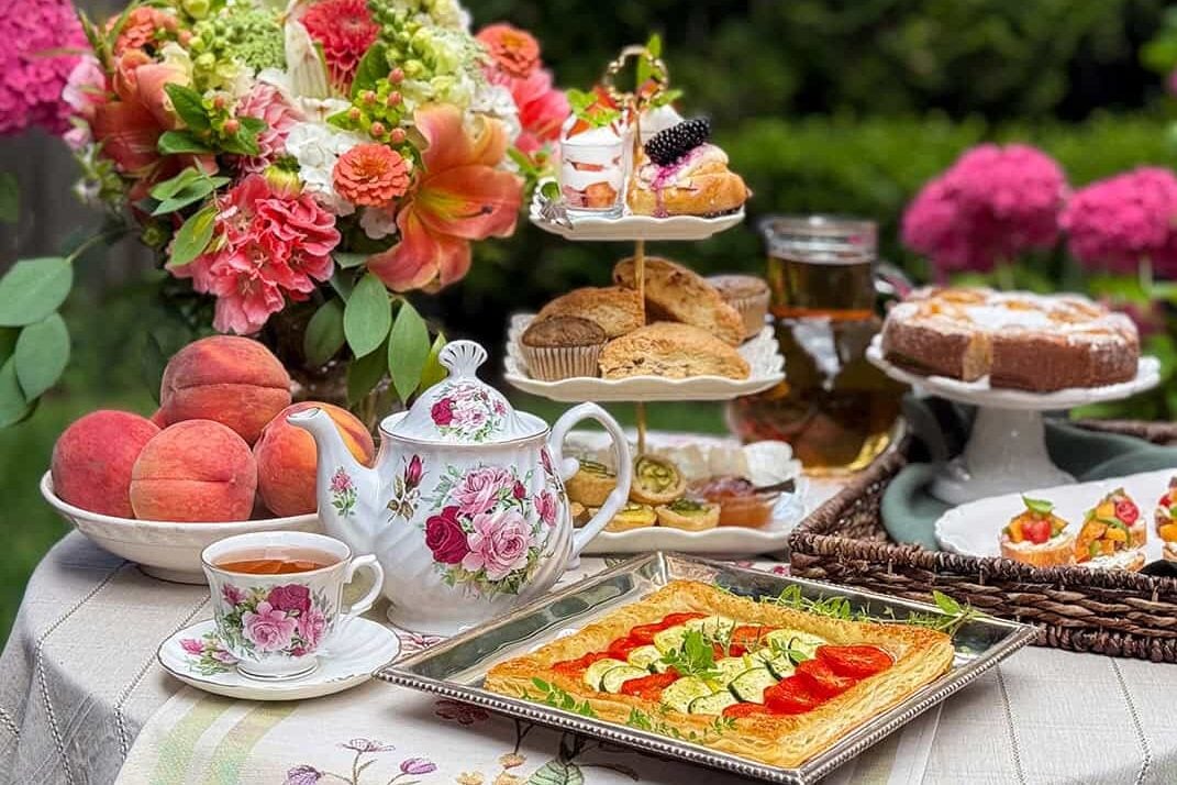 A charming August afternoon tea party is set outdoors with a floral teapot, cup of tea, peaches, a tiered tray of pastries, vegetable tart, cake, and flowers.