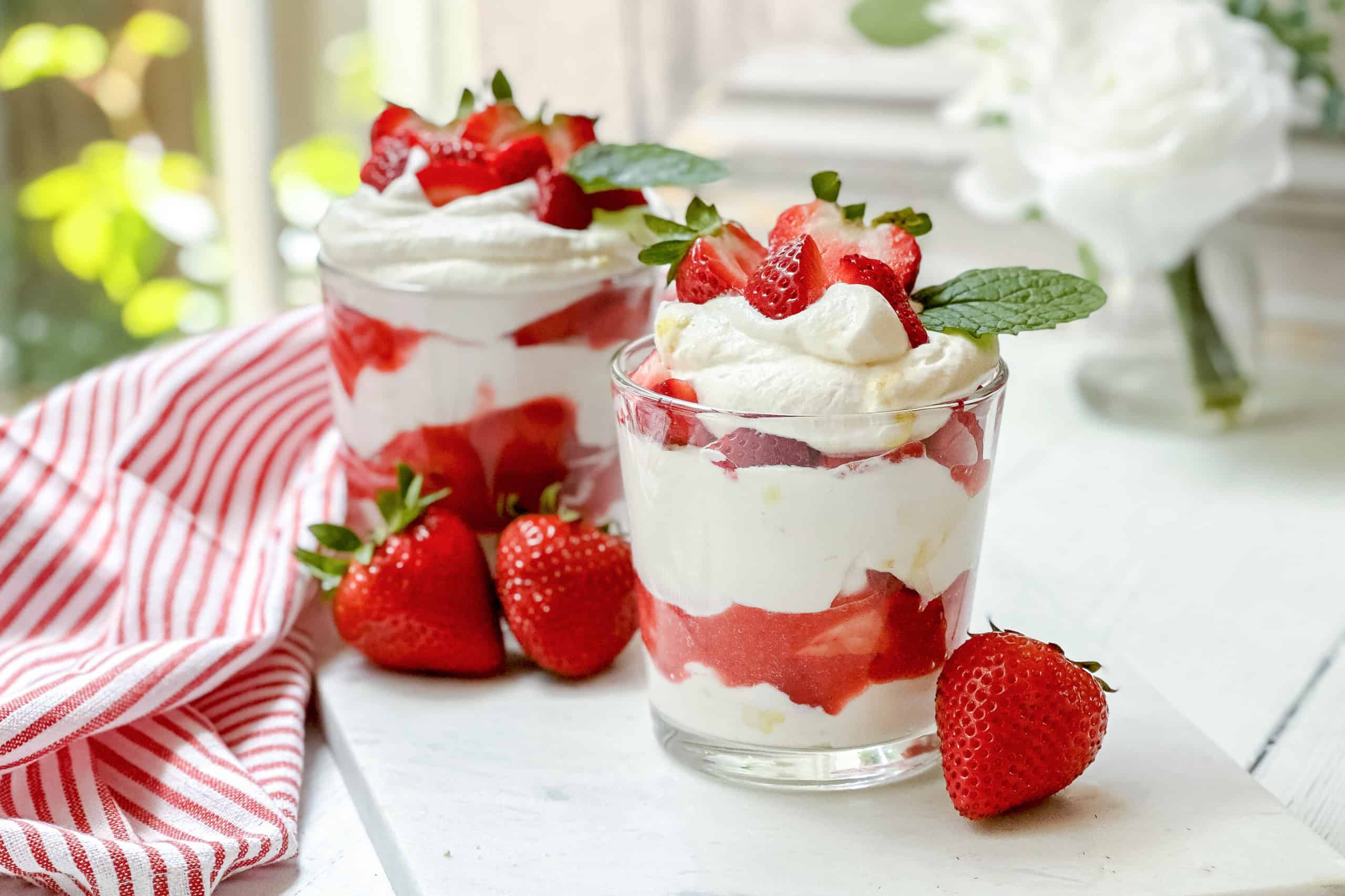 Two glasses filled with layers of whipped cream and strawberries, garnished with mint leaves, sit on a white surface beside fresh strawberries and a red-striped cloth-a perfect example of quick and easy recipes for a refreshing dessert.