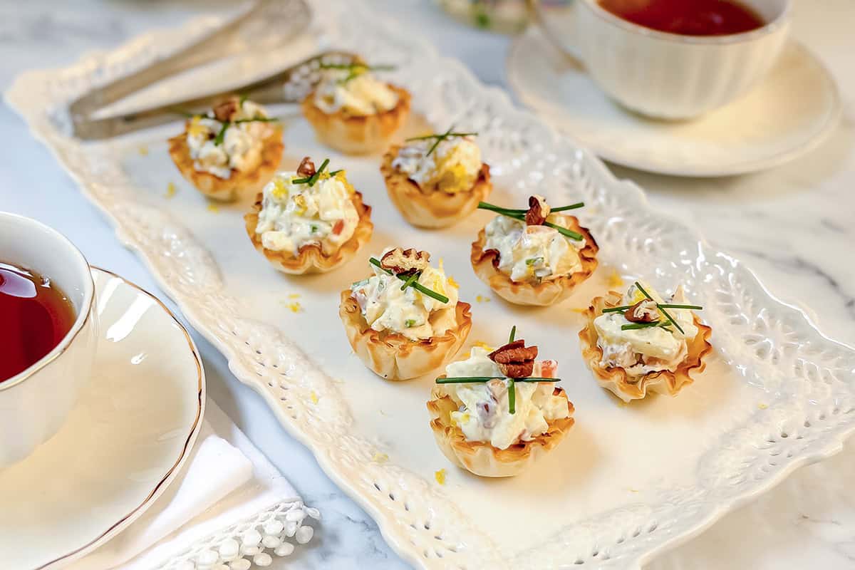 Mini phyllo tartlets filled with a creamy salad mixture, garnished with chopped herbs and pecans, arranged on a white serving platter next to a cup of tea.