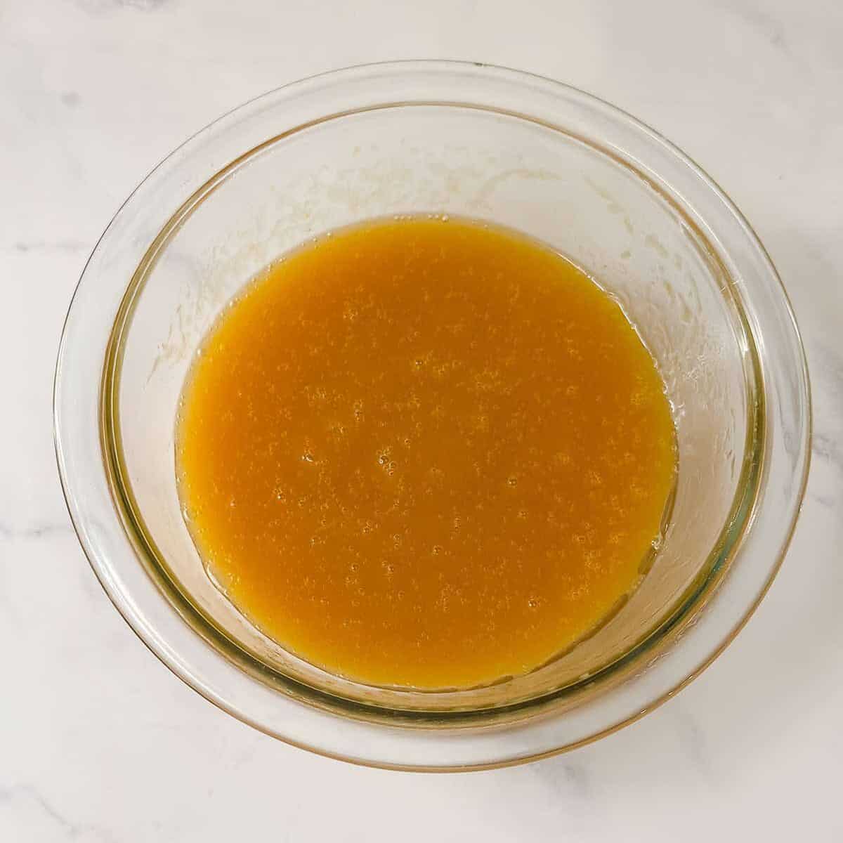 A clear glass bowl containing a thick orange-yellow liquid, perfect for baking muffins, sits on a white marble surface.