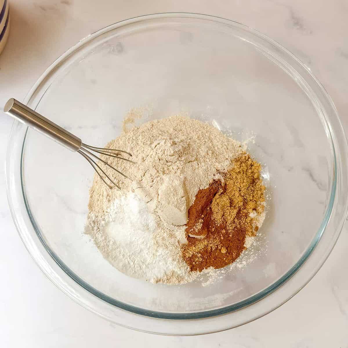A glass bowl with flour, seasonings, and leavening for these healthy zucchini muffins, plus a whisk, on a light marble countertop.