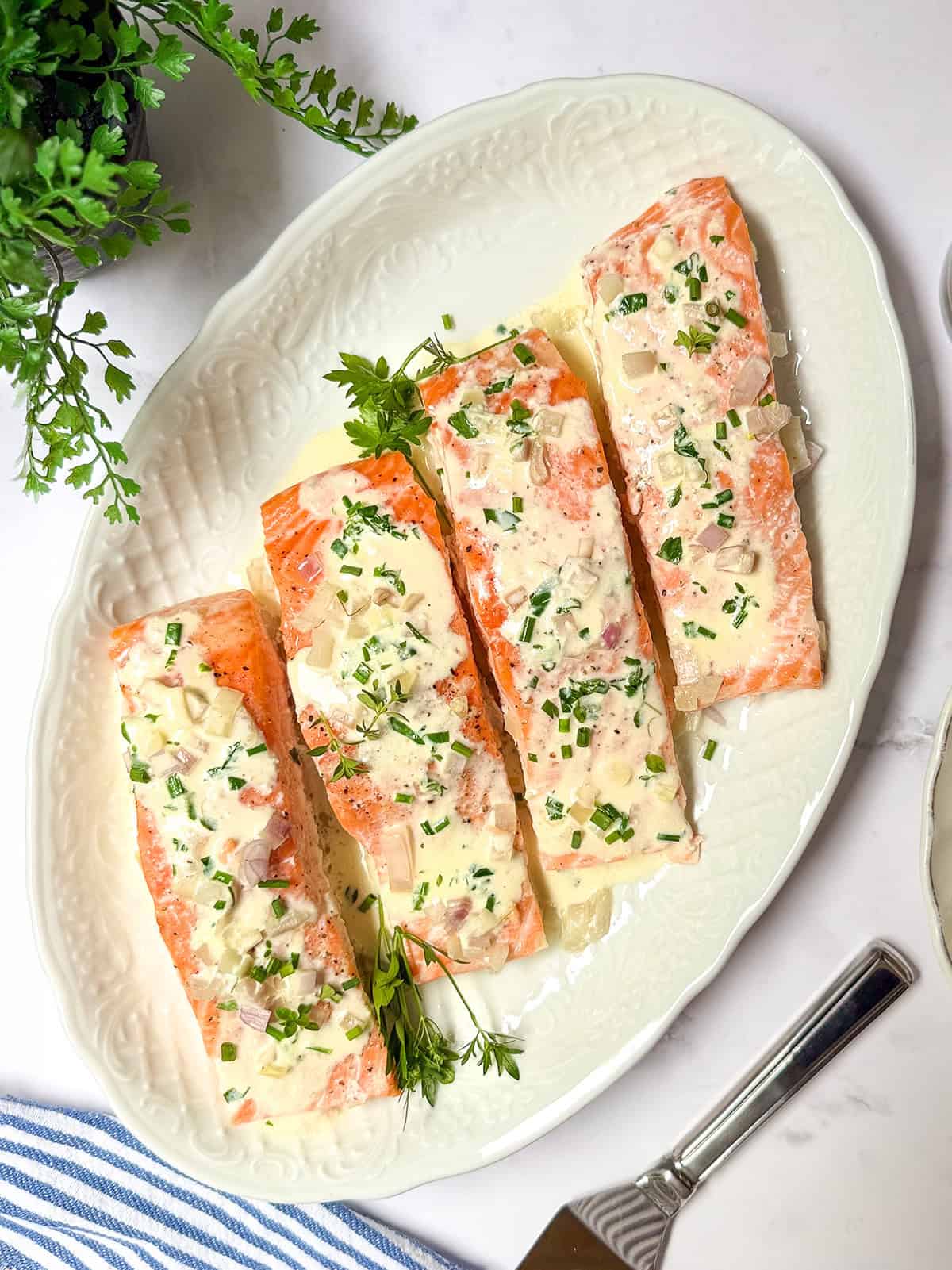 Four baked salmon fillets topped with a herb cream sauce are arranged on a white oval plate and garnished with fresh parsley.