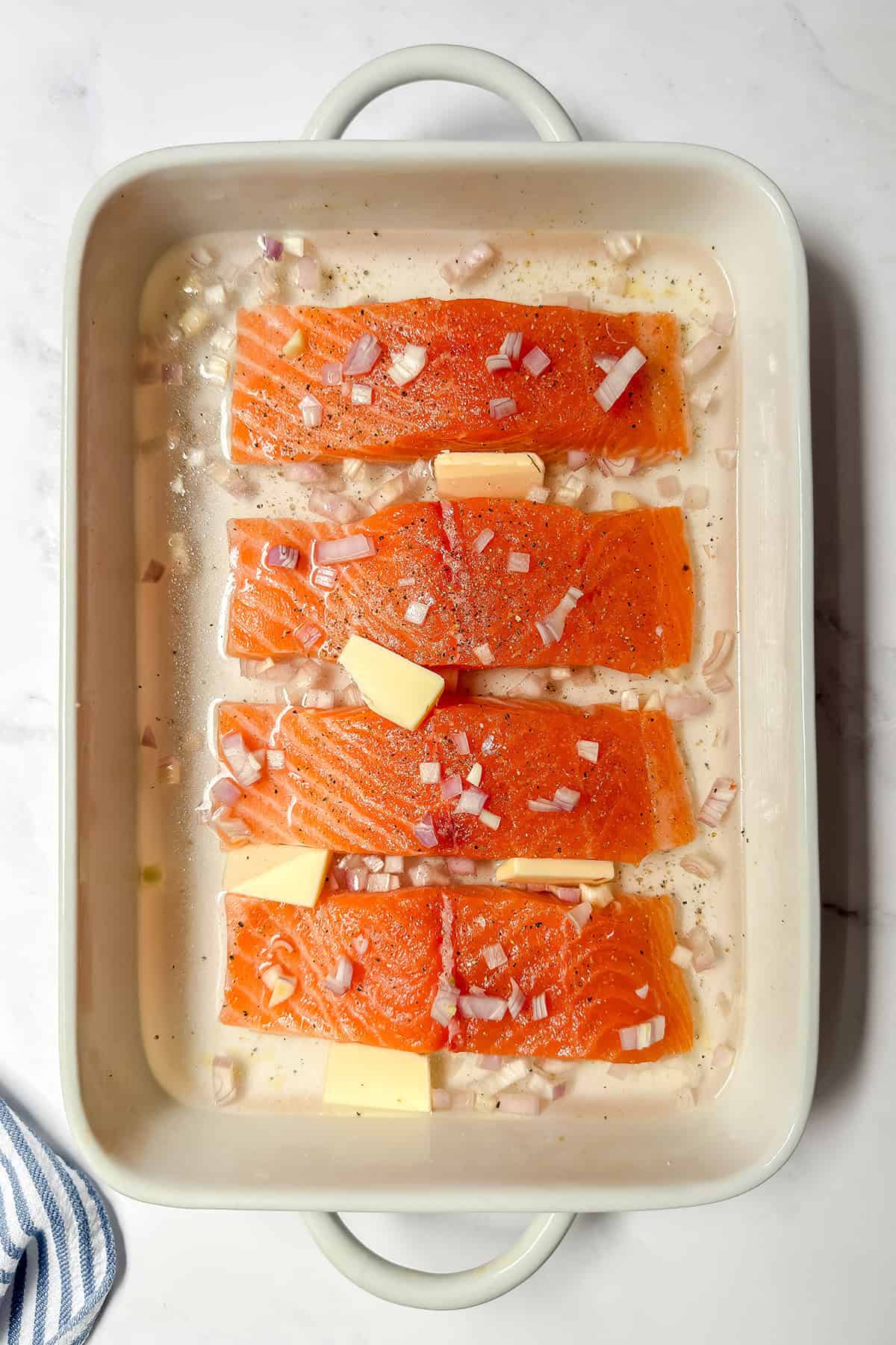 Four raw salmon fillets in a white baking dish, topped with chopped shallots and butter slices, ready to be baked.