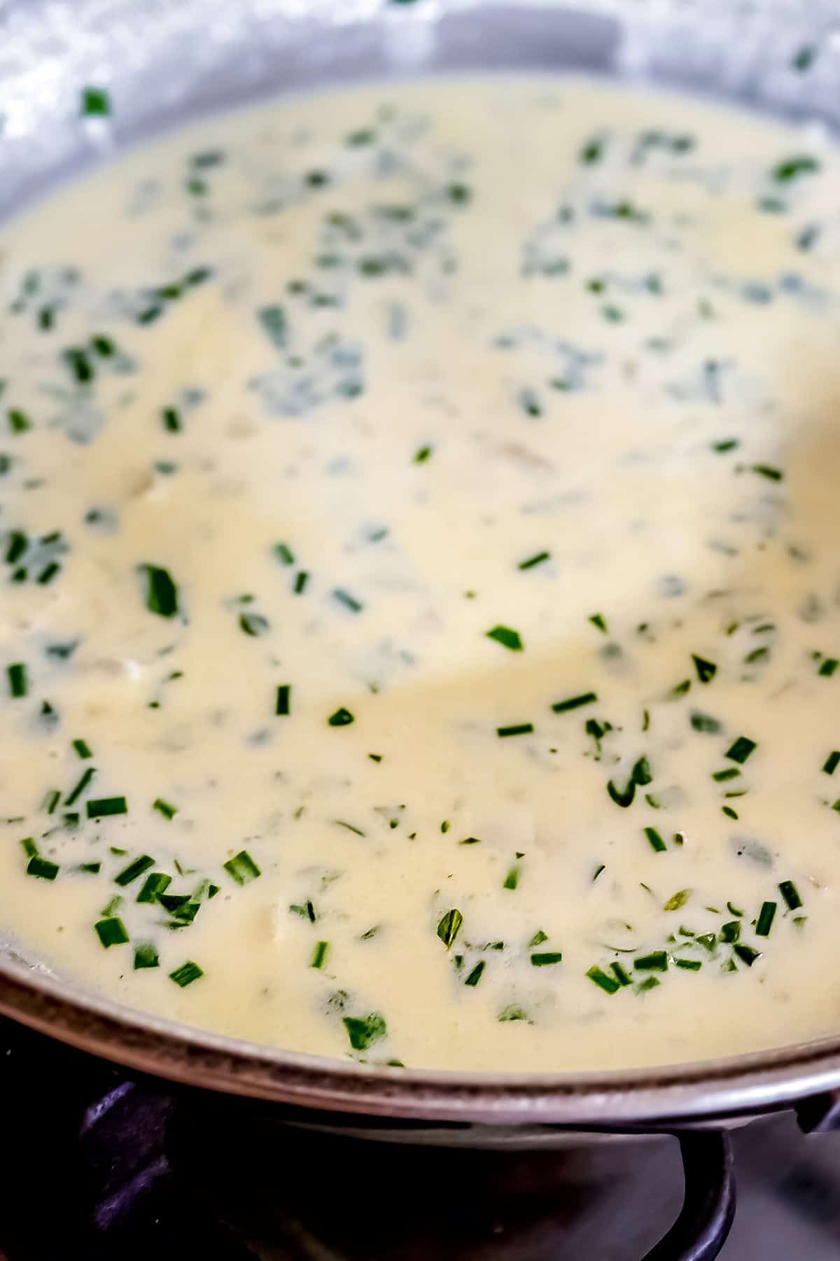 A close-up of a creamy herb cream sauce with chopped herbs simmering in a metal skillet on the stovetop.