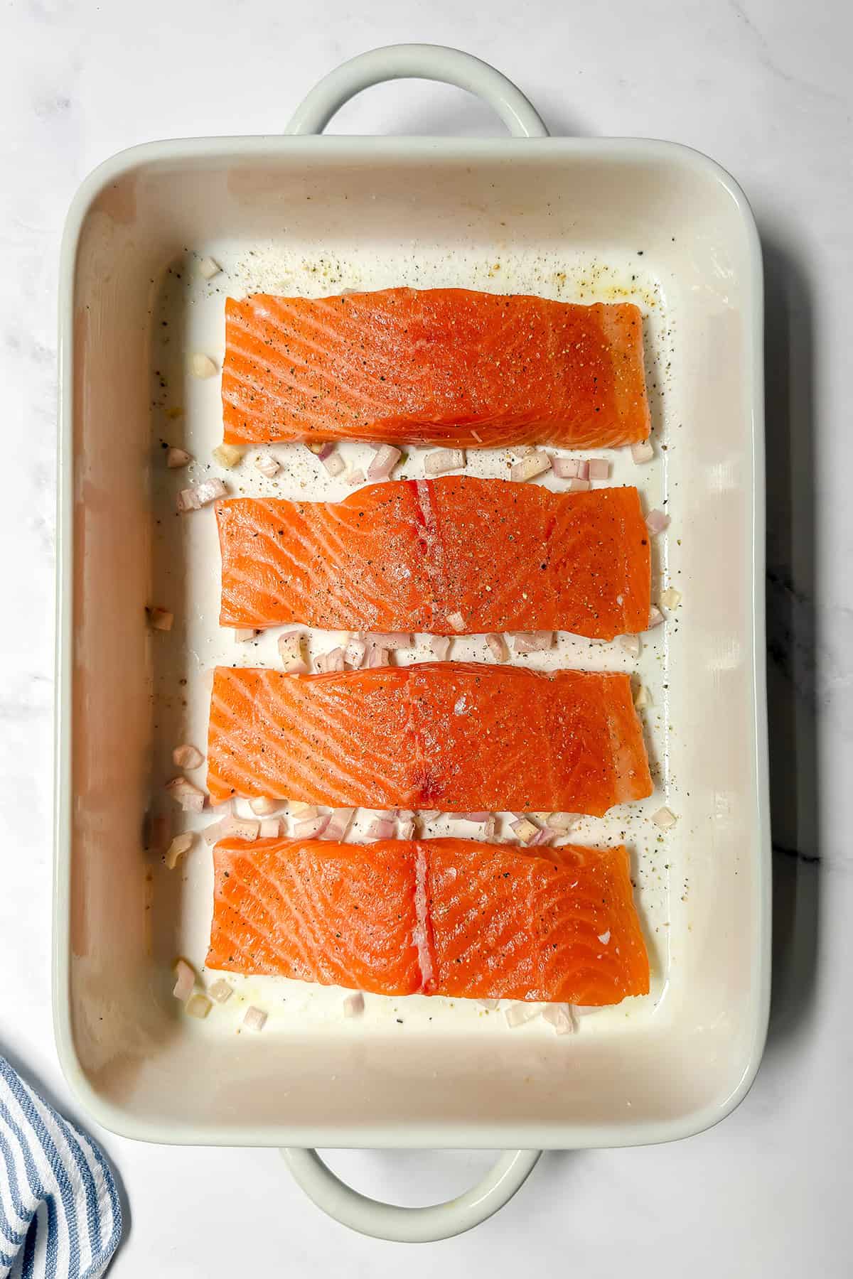 Four raw salmon fillets seasoned with pepper and surrounded by chopped onions are arranged in a white baking dish on a white surface.
