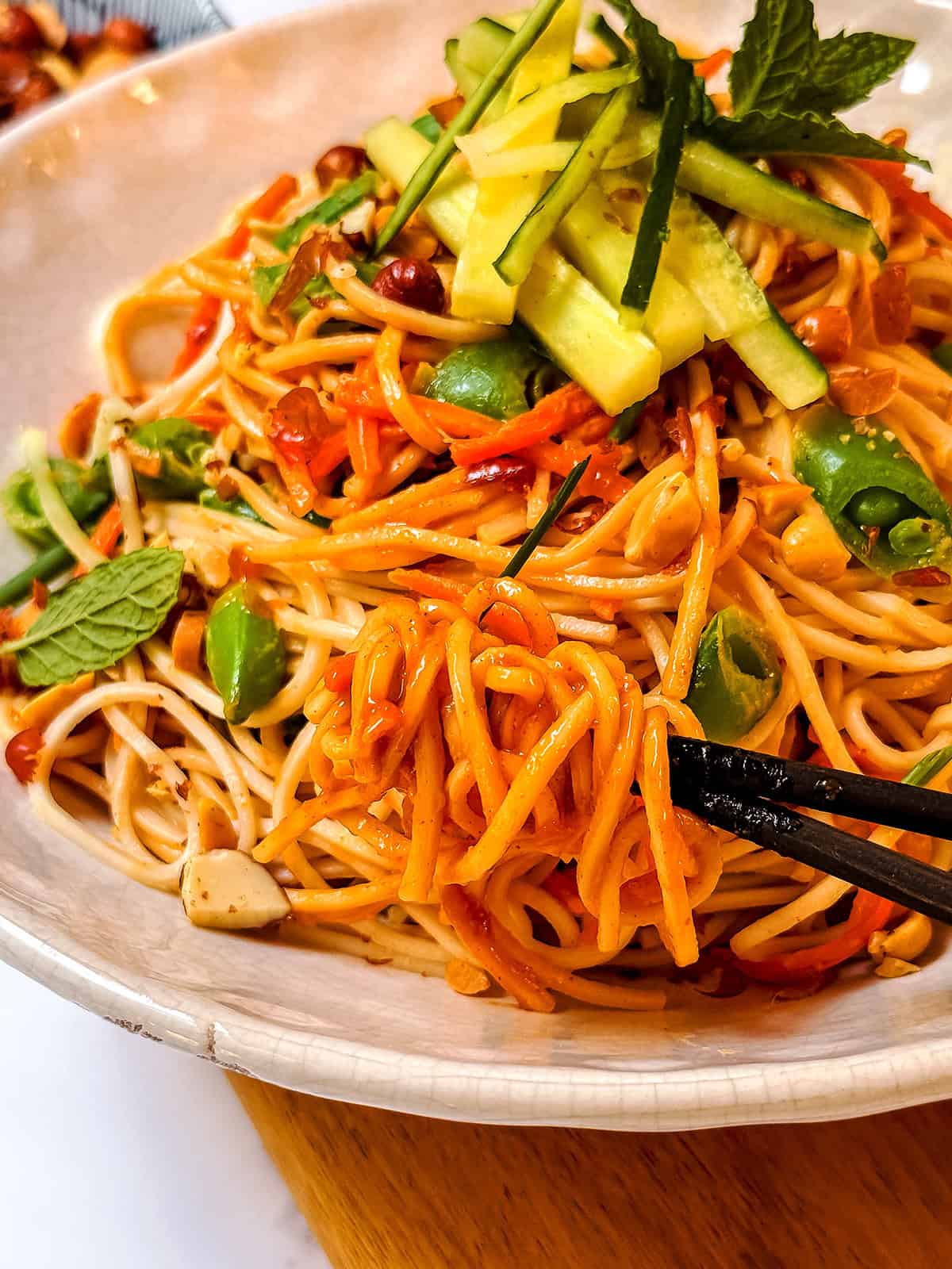 Chopsticks hold noodles from a bowl of Cold Peanut Sesame Noodles, an Asian-style pasta topped with cucumber strips, carrots, edamame, peanuts, and mint.