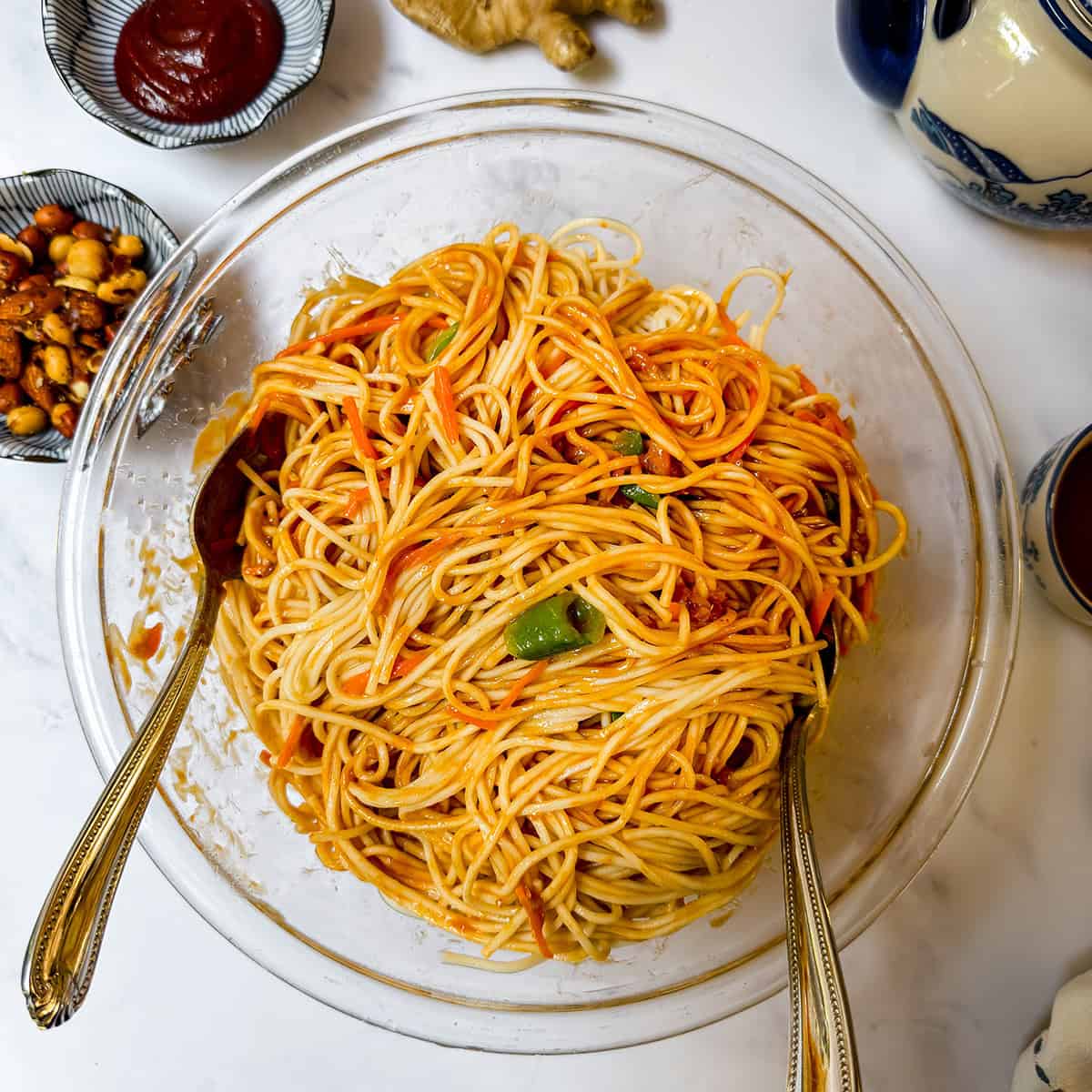 A glass bowl of saucy spaghetti with vegetables sits on a marble surface, reminiscent of Cold Peanut Sesame Noodles, surrounded by small dishes of sauce and nuts, two spoons placed in the pasta.