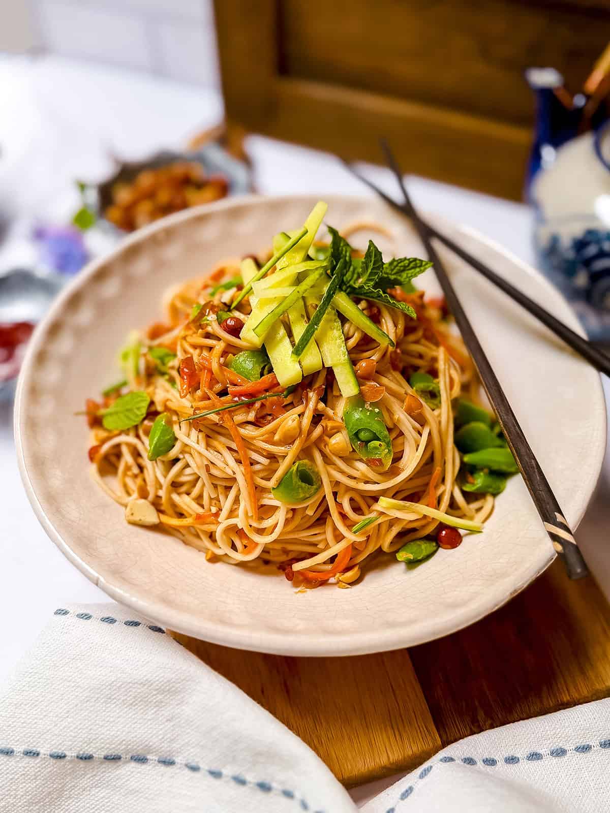 A plate of Cold Peanut Sesame Noodles garnished with sliced cucumber, vegetables, and herbs, with chopsticks resting on the rim of the bowl.