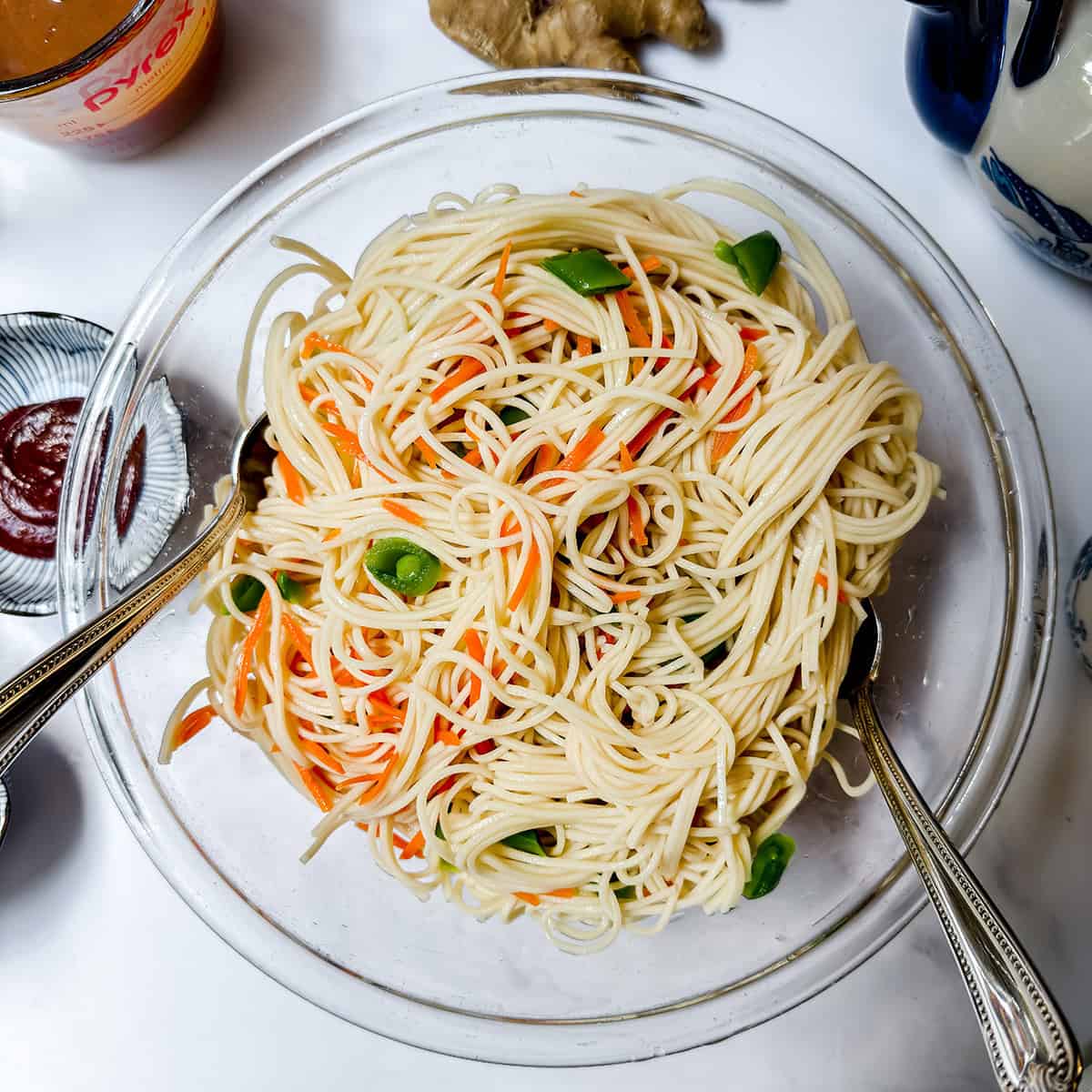 A clear glass bowl filled with Cold Peanut Sesame Noodles mixed with sliced carrots and green vegetables, with two metal serving spoons in the bowl, on a white surface.