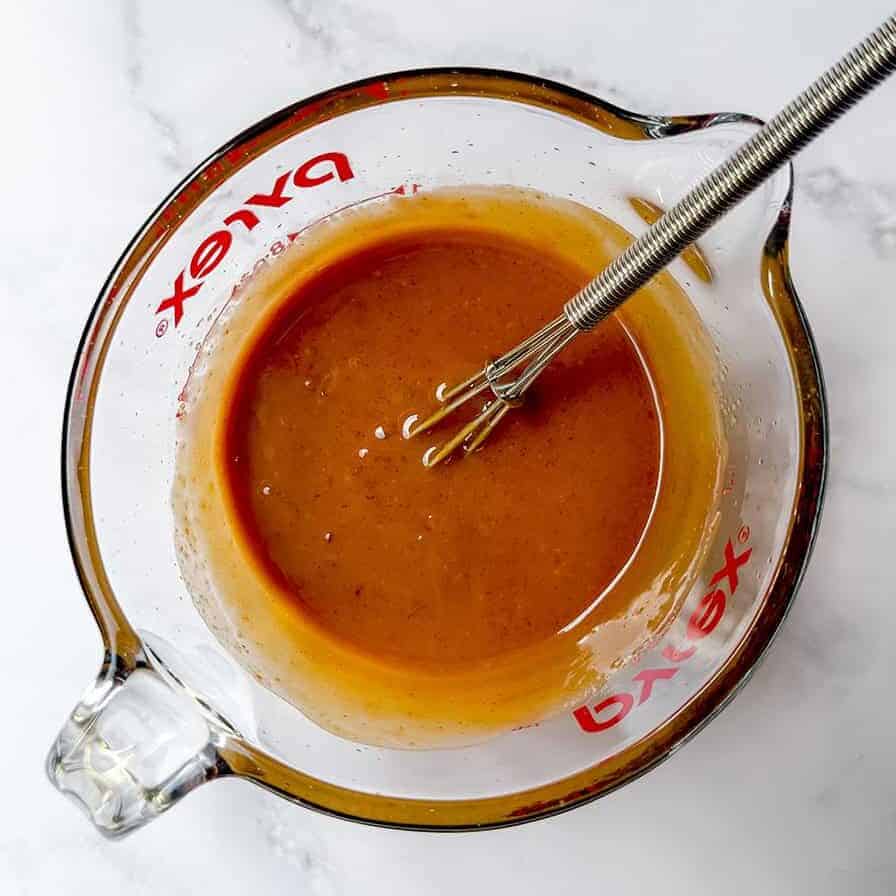 Glass measuring cup with light brown sauce being whisked on a white marble surface, perfect for drizzling over Cold Peanut Sesame Noodles.