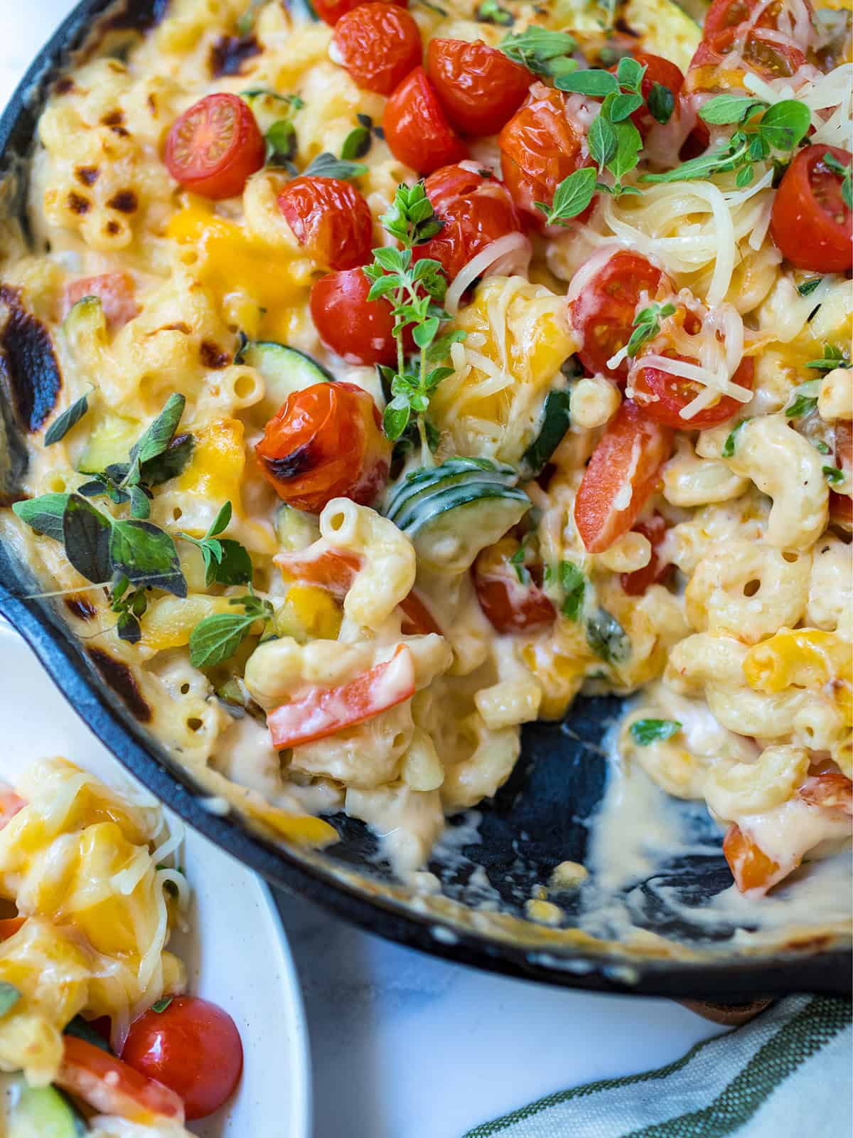 A skillet of baked mac and cheese mixed with fresh tomatoes, zucchini, savory herbs, and melted cheese, with a generous serving of this skillet mac and cheese scooped onto a plate.