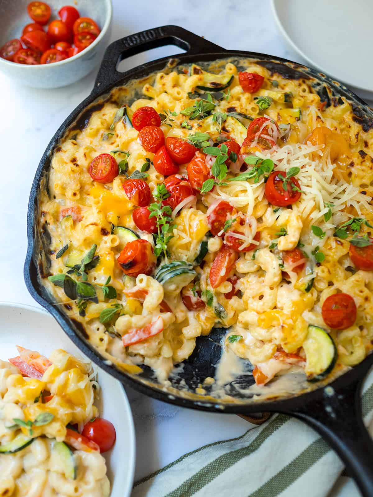 A cast iron skillet brimming with creamy Skillet Mac and Cheese, generously topped with cherry tomatoes, zucchini, shredded cheese, and fresh herbs. Nearby, a bowl of tomatoes and a plated serving invite you to enjoy this comforting dish.