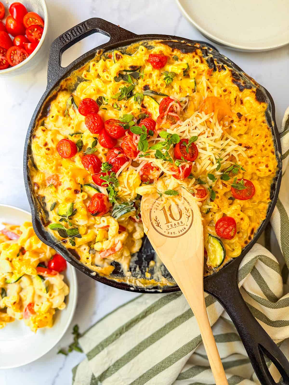 A cast iron skillet filled with baked mac and cheese topped with cherry tomatoes, zucchini, herbs, and shredded cheese, next to a wooden spoon and a bowl of the dish.