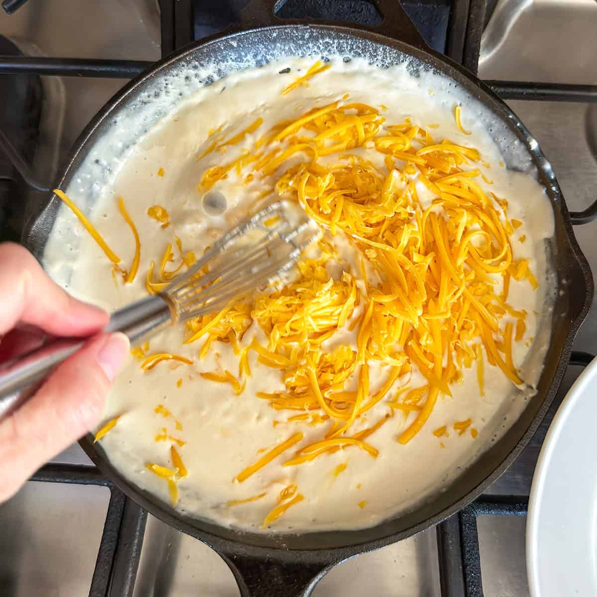 A whisk stirs shredded cheddar cheese into a creamy white sauce in a skillet on a stovetop.