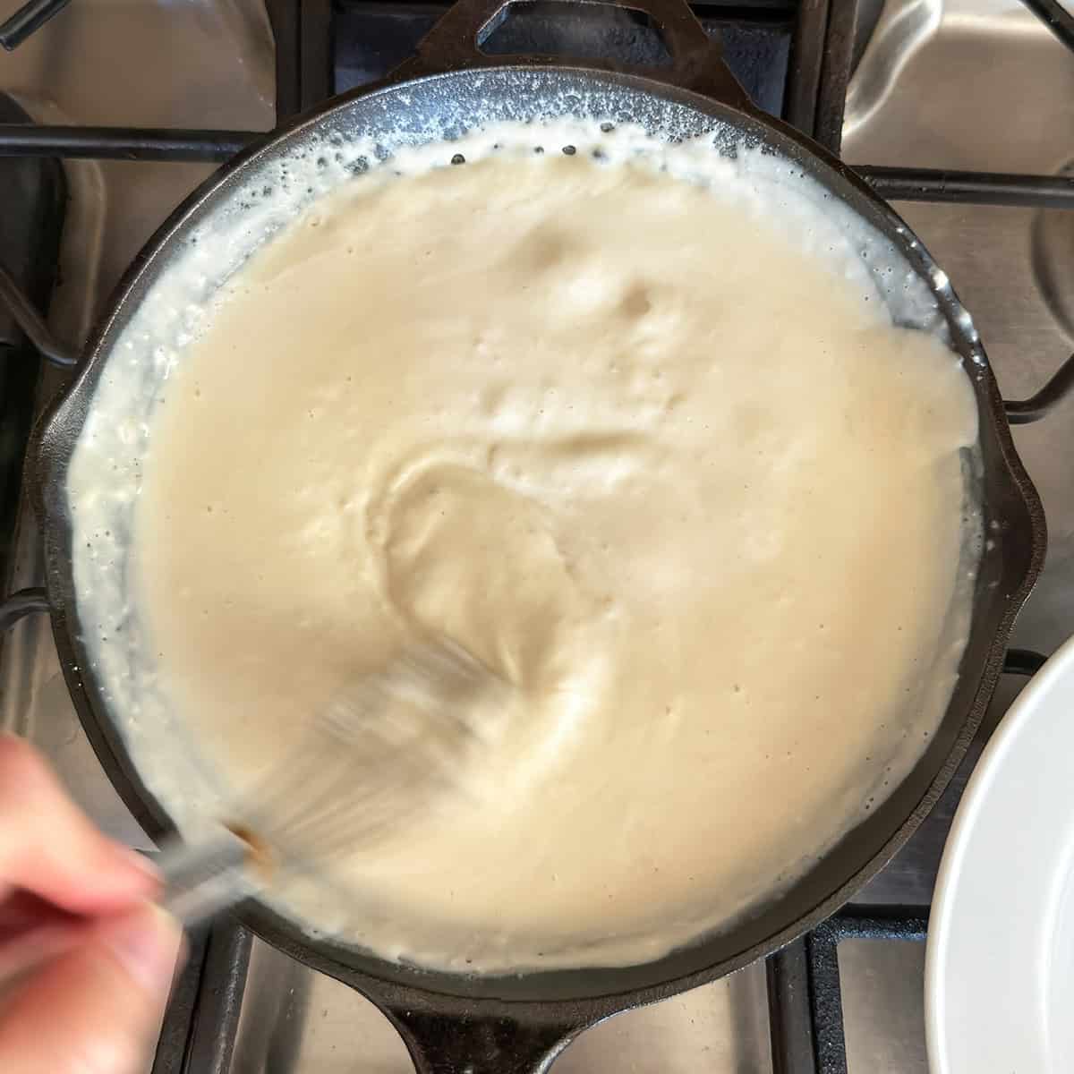 A hand whisks a thick, creamy white sauce in a black skillet on a stovetop.