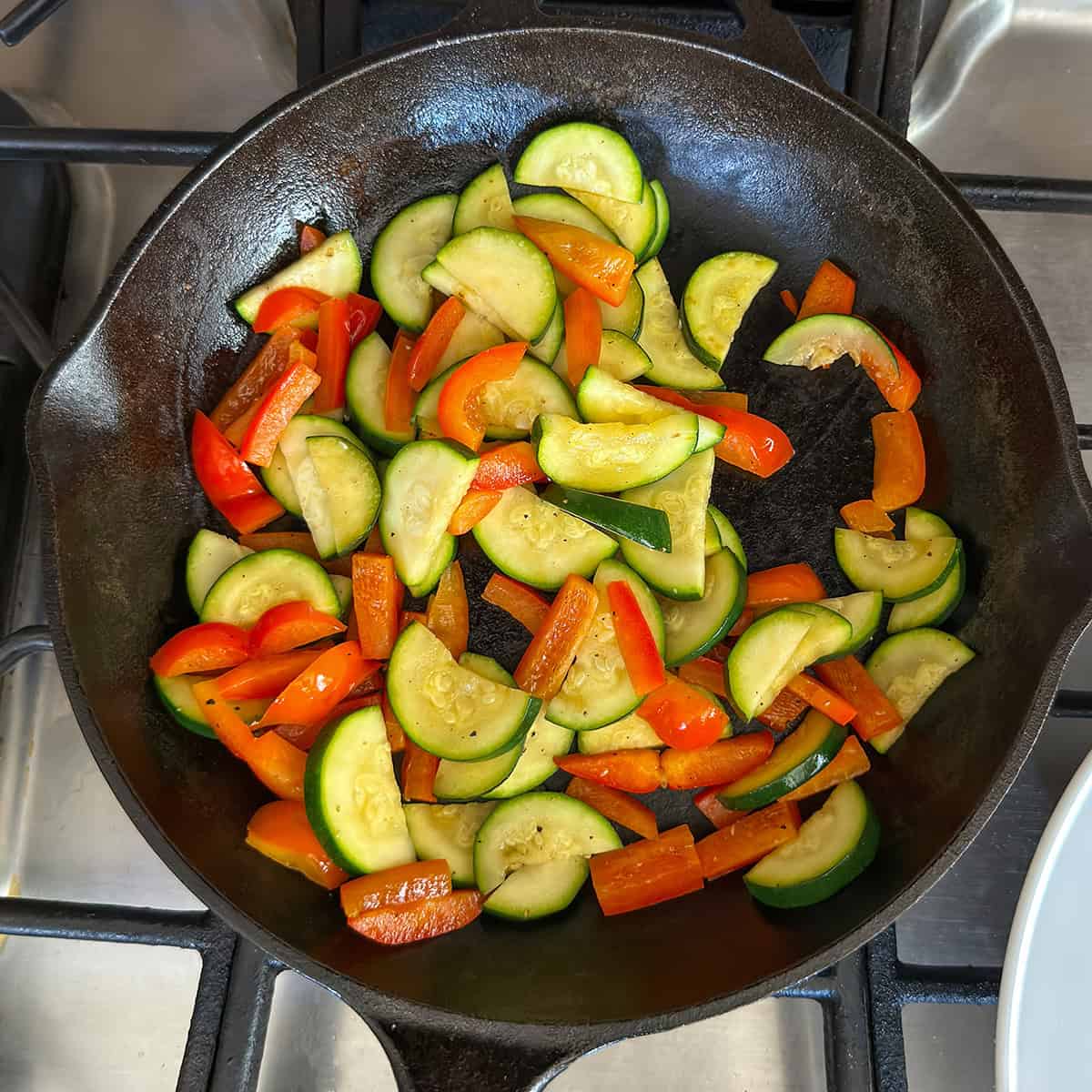 Sliced zucchini and red bell peppers sauté in a black cast iron skillet on a stovetop.