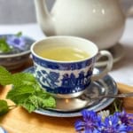 A cup of light-colored Liberty Tea in a blue and white patterned cup sits on a matching saucer with a spoon, surrounded by fresh mint leaves, fresh herbs, and purple flowers, with a teapot in the background.
