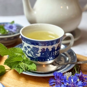 A cup of light-colored Liberty Tea in a blue and white patterned cup sits on a matching saucer with a spoon, surrounded by fresh mint leaves, fresh herbs, and purple flowers, with a teapot in the background.