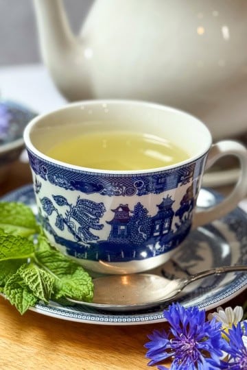 A cup of light-colored Liberty Tea in a blue and white patterned cup sits on a matching saucer with a spoon, surrounded by fresh mint leaves, fresh herbs, and purple flowers, with a teapot in the background.
