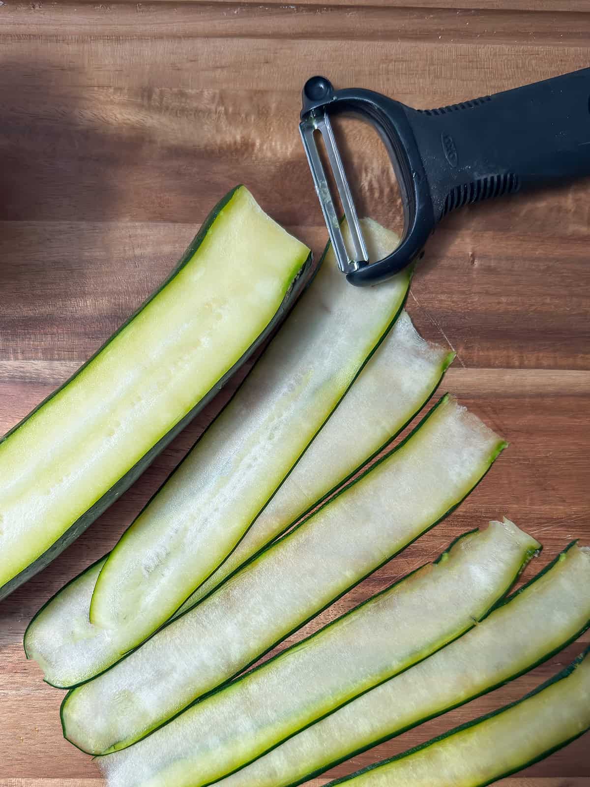 Thin zucchini ribbons beside a wide vegetable peeler on a wooden cutting board.