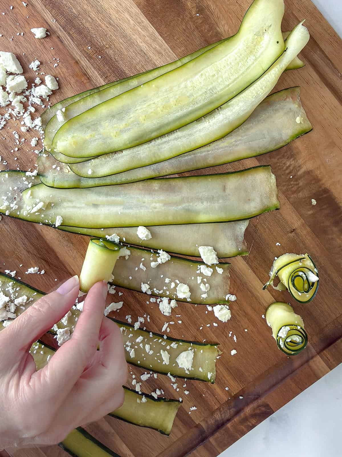 A hand rolls thin zucchini slices with crumbled feta cheese on a wooden cutting board, with finished zucchini rolls.