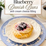 A Cream Cheese Danish bun filled with sweet blueberry and topped with icing sits on a white plate with a blue floral border; coffee and more buns are in the background.