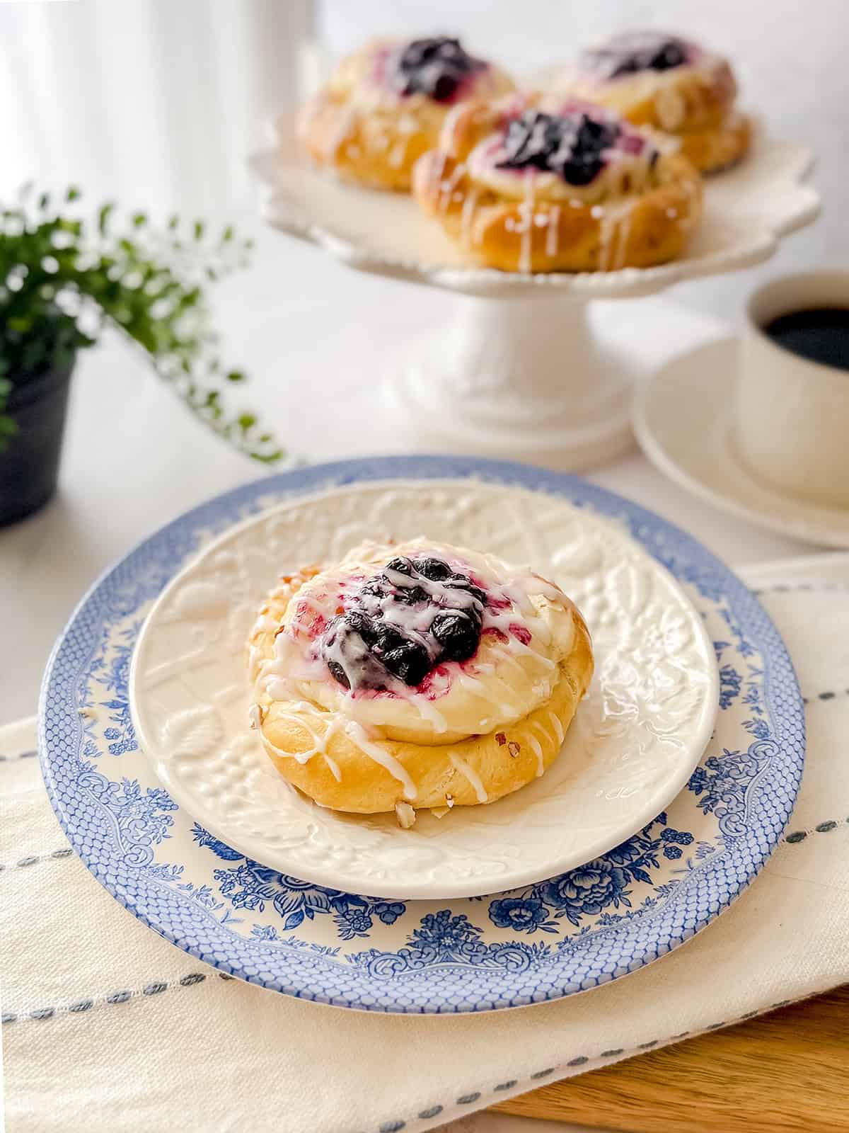 A Danish Bun topped with blueberry filling and icing sits on a patterned plate, with more pastries on a cake stand and a cup of coffee in the background.