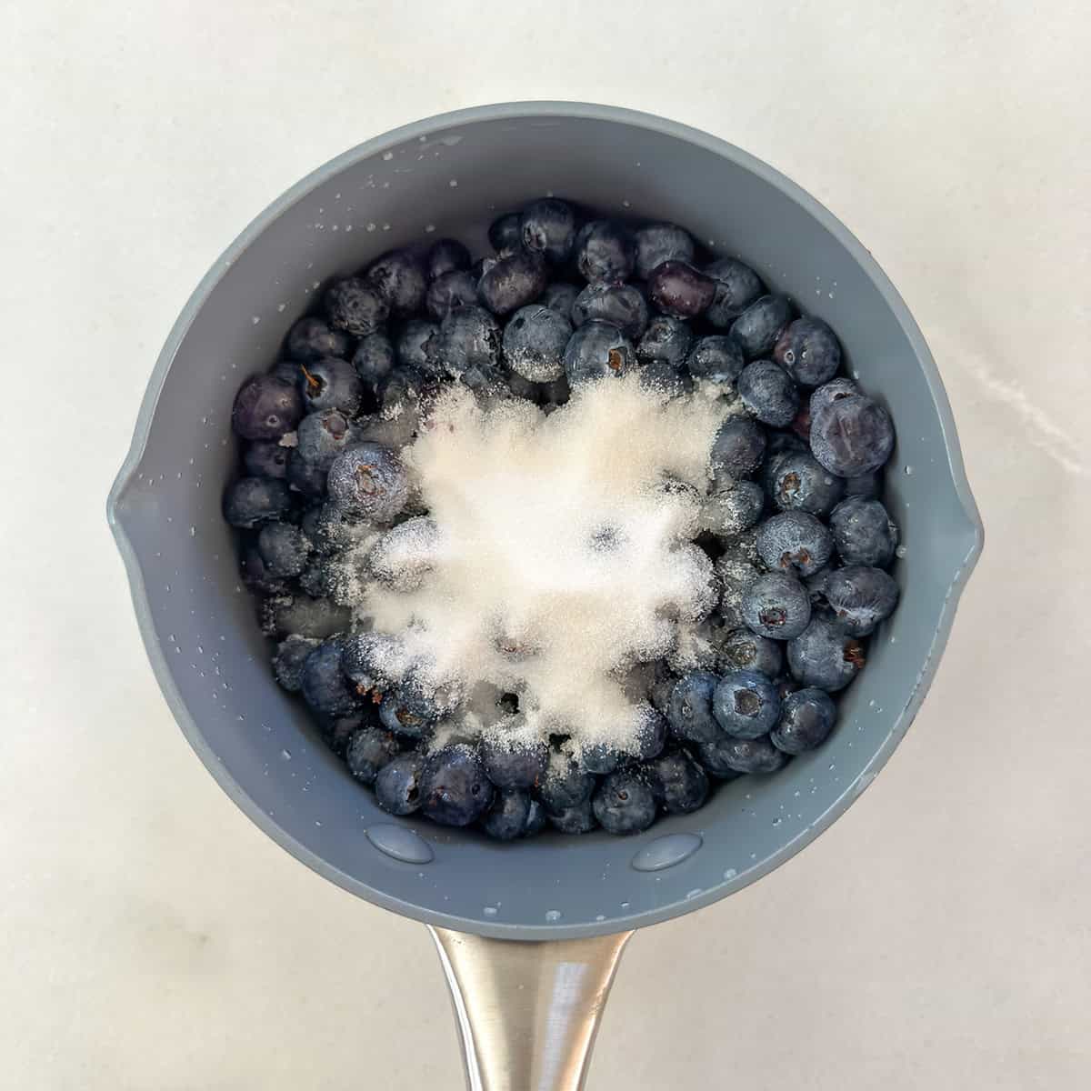 A gray saucepan filled with fresh blueberries, destined for a classic Blueberry Danish, is topped with a mound of granulated white sugar and sits on a white surface.