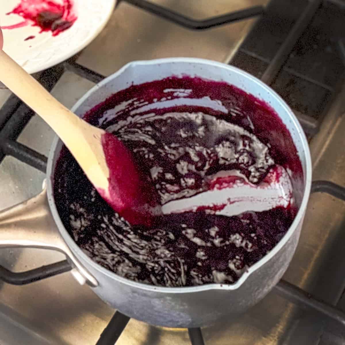 A saucepan containing a thick, dark purple mixture-perfect for filling a Blueberry Danish-is being stirred with a wooden spoon on a stovetop.