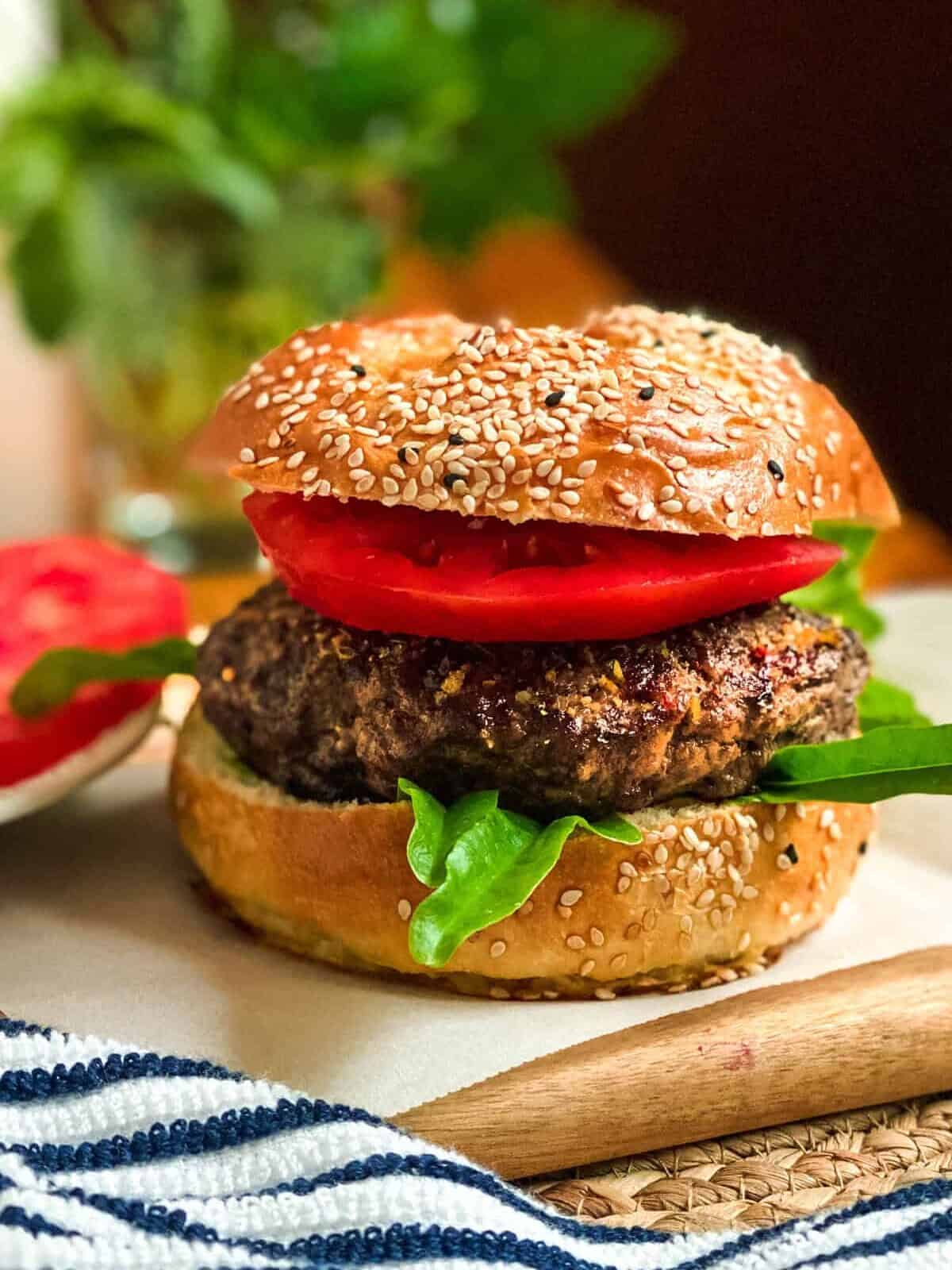 A close-up of a sesame seed bun burger with a thick patty, fresh spinach leaves, and sliced tomato, perfect for any burgers recipe, with blurred greens and a striped napkin in the background.