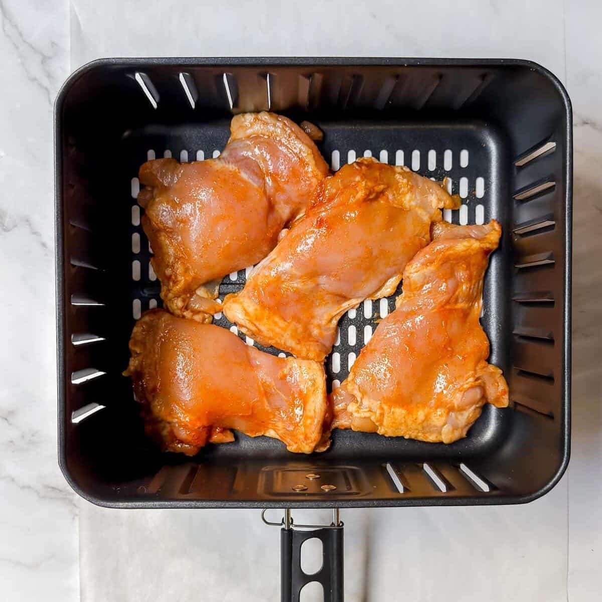 Raw chicken thighs, destined for delicious chicken tacos, are seasoned with a reddish marinade and arranged in a black air fryer basket on a marble countertop.
