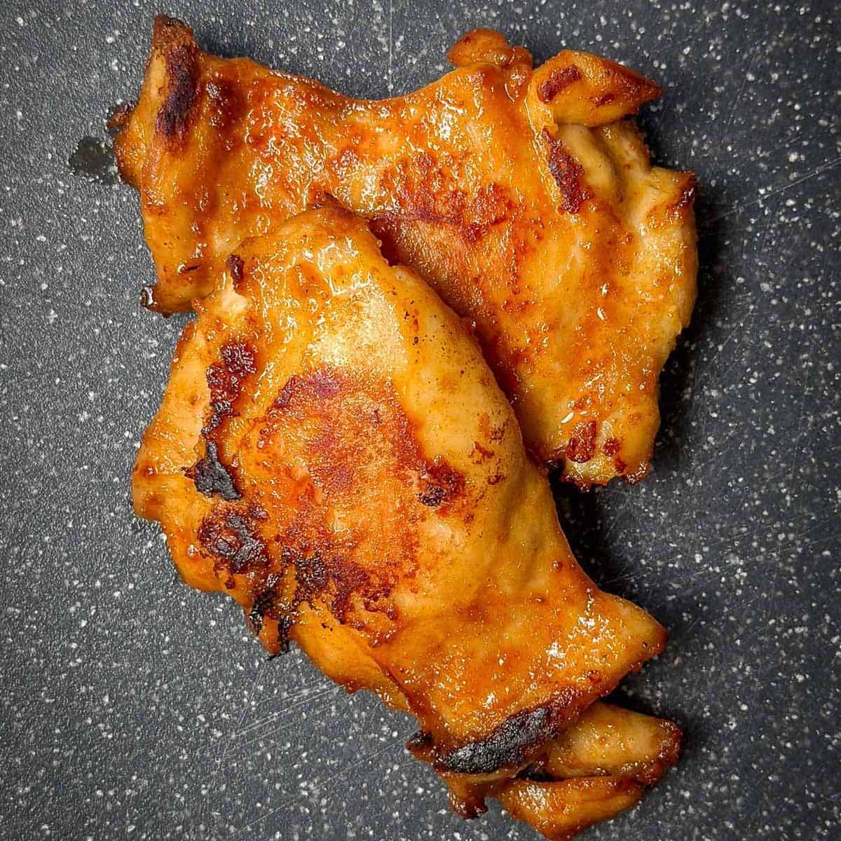 Two pieces of grilled chicken with a golden-brown crust rest on a black speckled surface, embodying the perfect filling for delectable chicken tacos. Their well-cooked exterior promises flavor in every bite.
