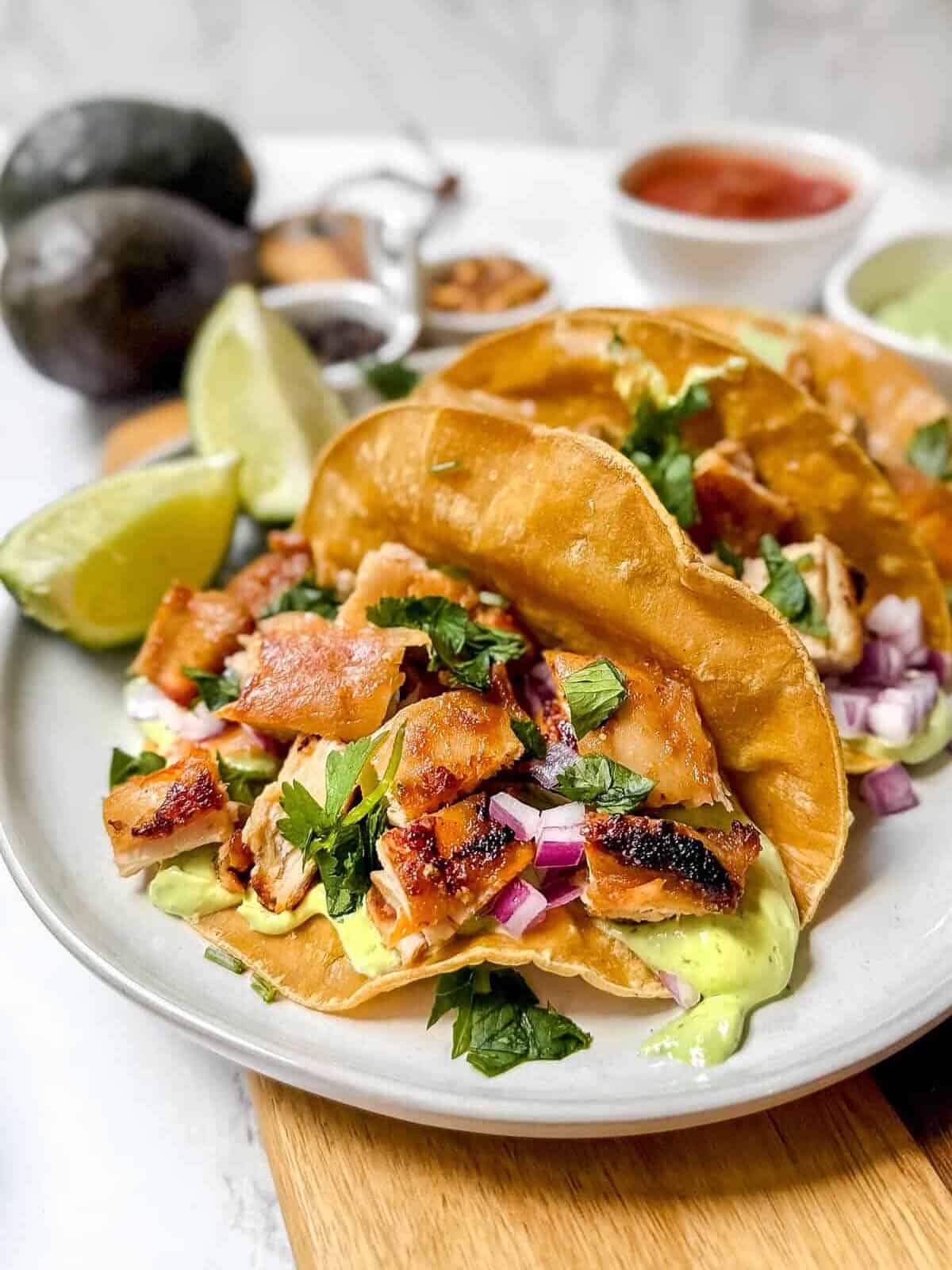 A plate with two tacos filled with grilled chicken, diced red onions, and cilantro, drizzled with green sauce. Lime wedges are on the side. Avocados and a bowl of salsa are in the blurred background.