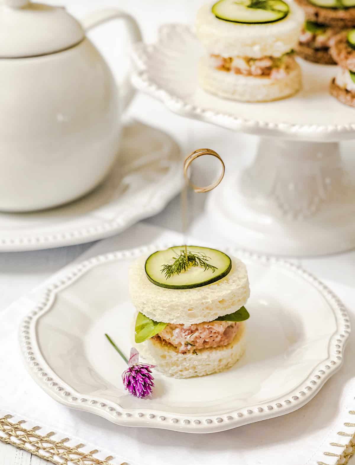 Round ham salad tea sandwich topped with cucumber and dill on a white plate, with a teapot and additional sandwiches in the background.