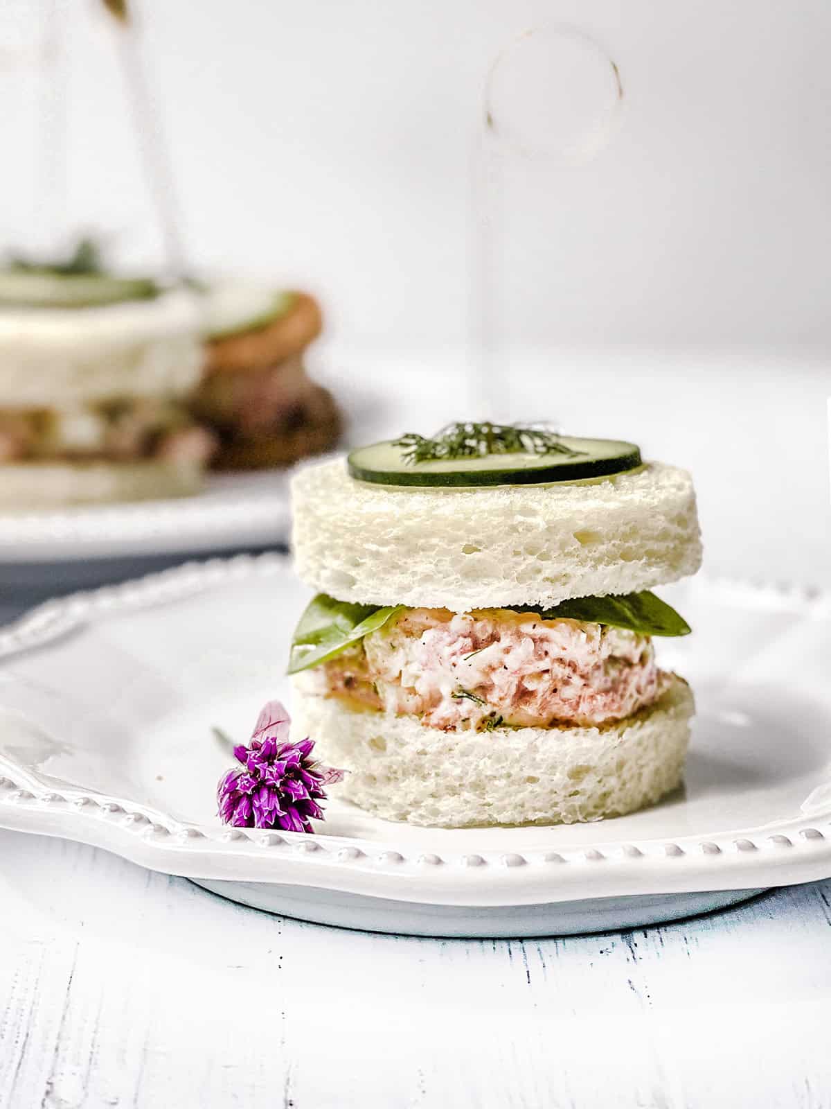 Close-up of a round ham salad tea sandwich with greens, cucumber, and fresh dill on a white plate.