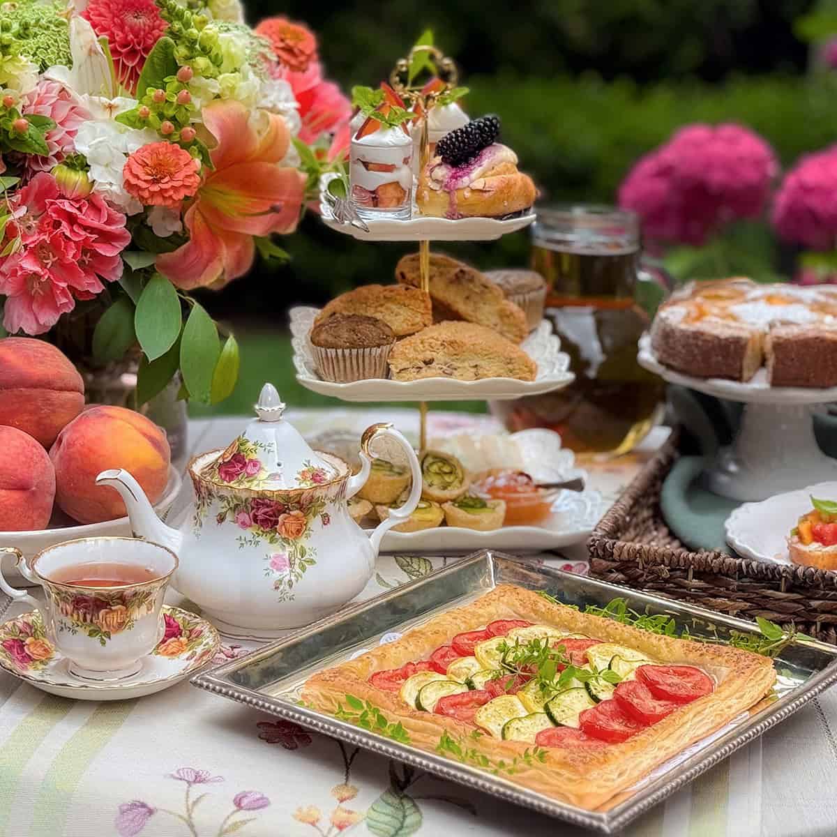 A table set outdoors with floral china tea set, a tray of vegetable tart made from quick and easy recipes, a tiered tray of pastries, fruit, cake slices, and fresh flowers.