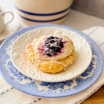 A Danish bun topped with white icing and a luscious blueberry filling sits on a white plate with a blue floral border, placed on a white napkin.